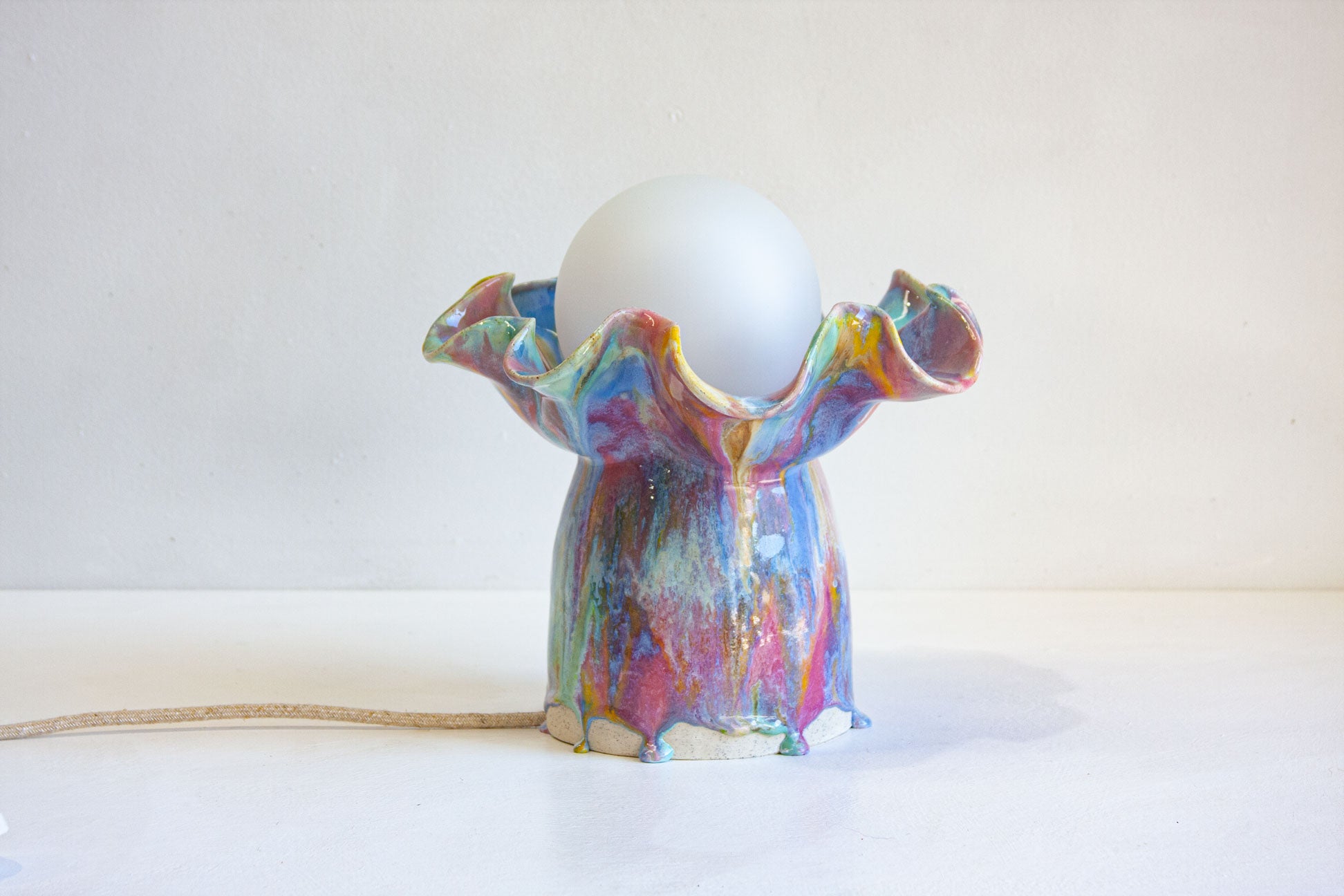 A handmade ceramic table lamp with a ruffled, dripping multicolor glaze and a frosted white glass globe shade, set against a plain off-white background.