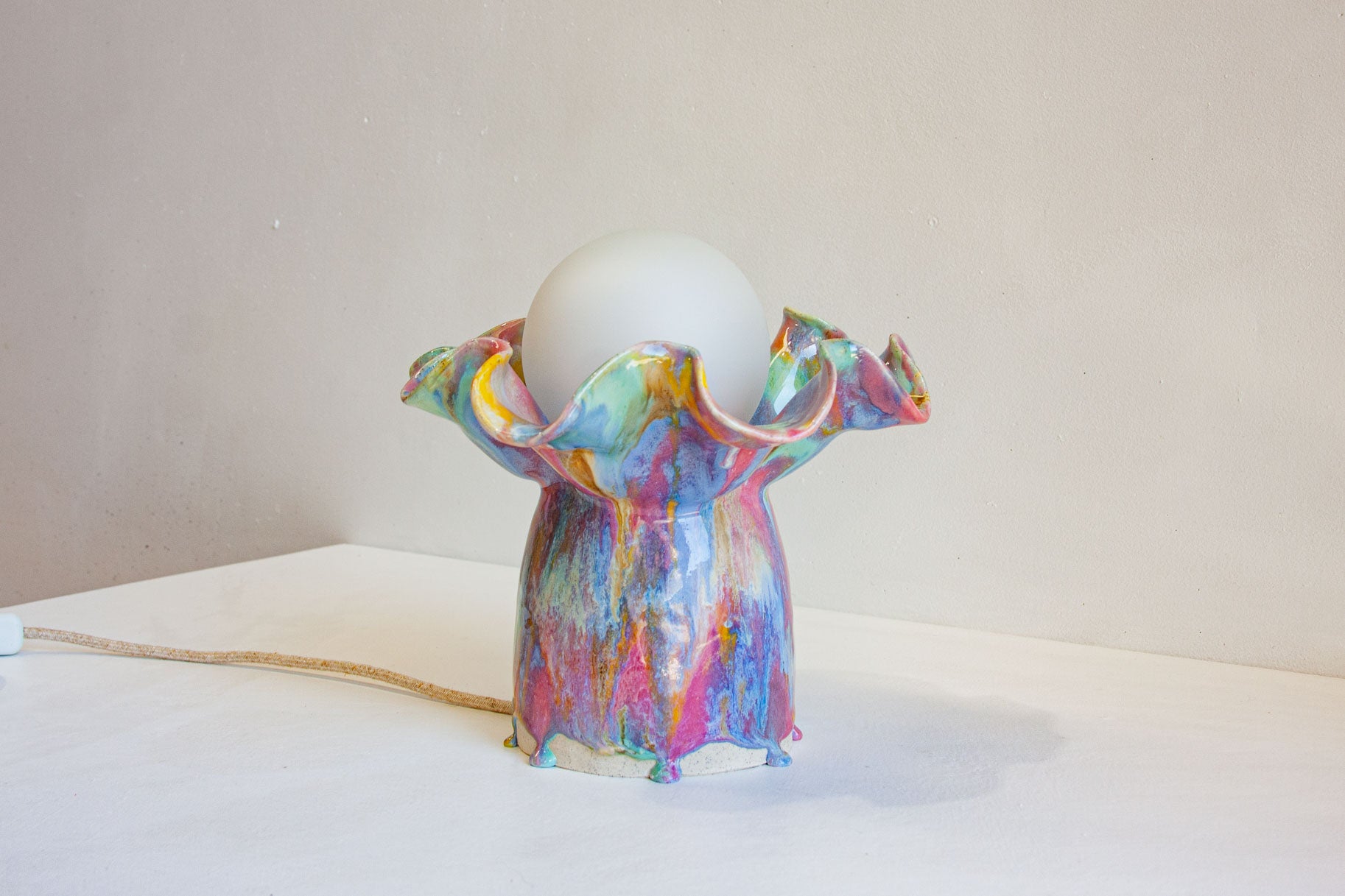 A colorful, sculptural ceramic lamp base with a white globe shade on top, set on a white surface against a plain beige wall.