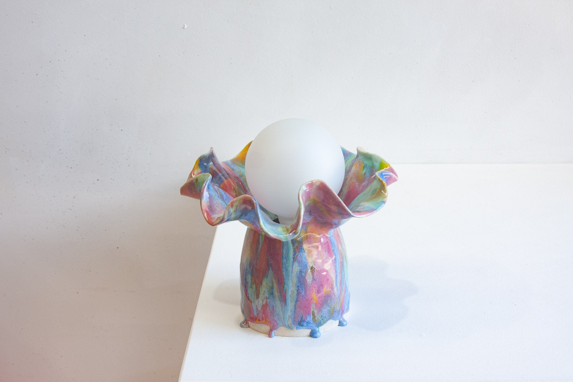 A colorful, sculptural ceramic lamp holder with a white spherical shade, displayed on a white surface against a light gray background.