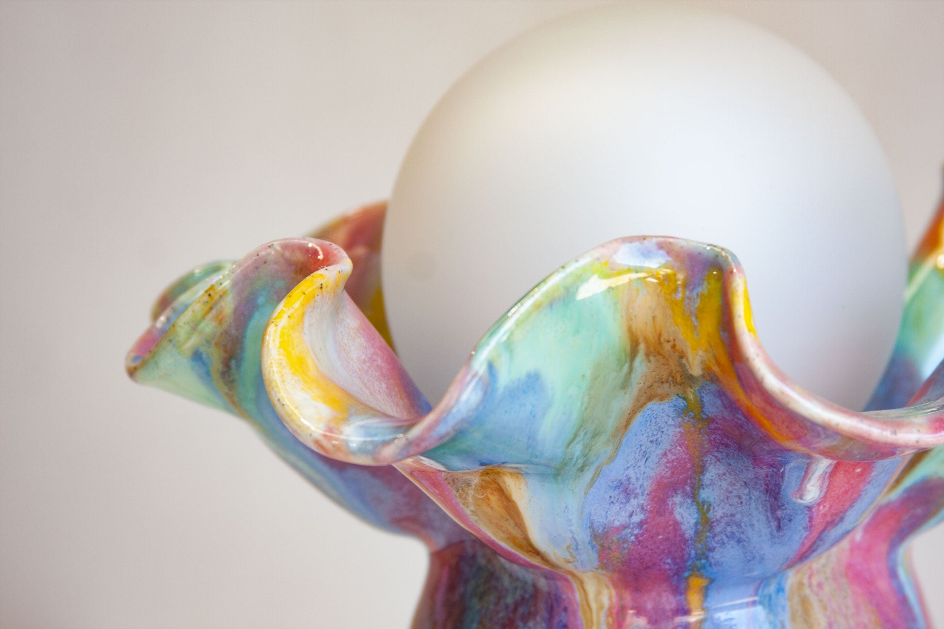 A close-up of a colorful, fluted ceramic vase or candle holder with a glossy marbled finish in shades of blue, pink, purple, yellow, and green, holding a smooth white spherical object.