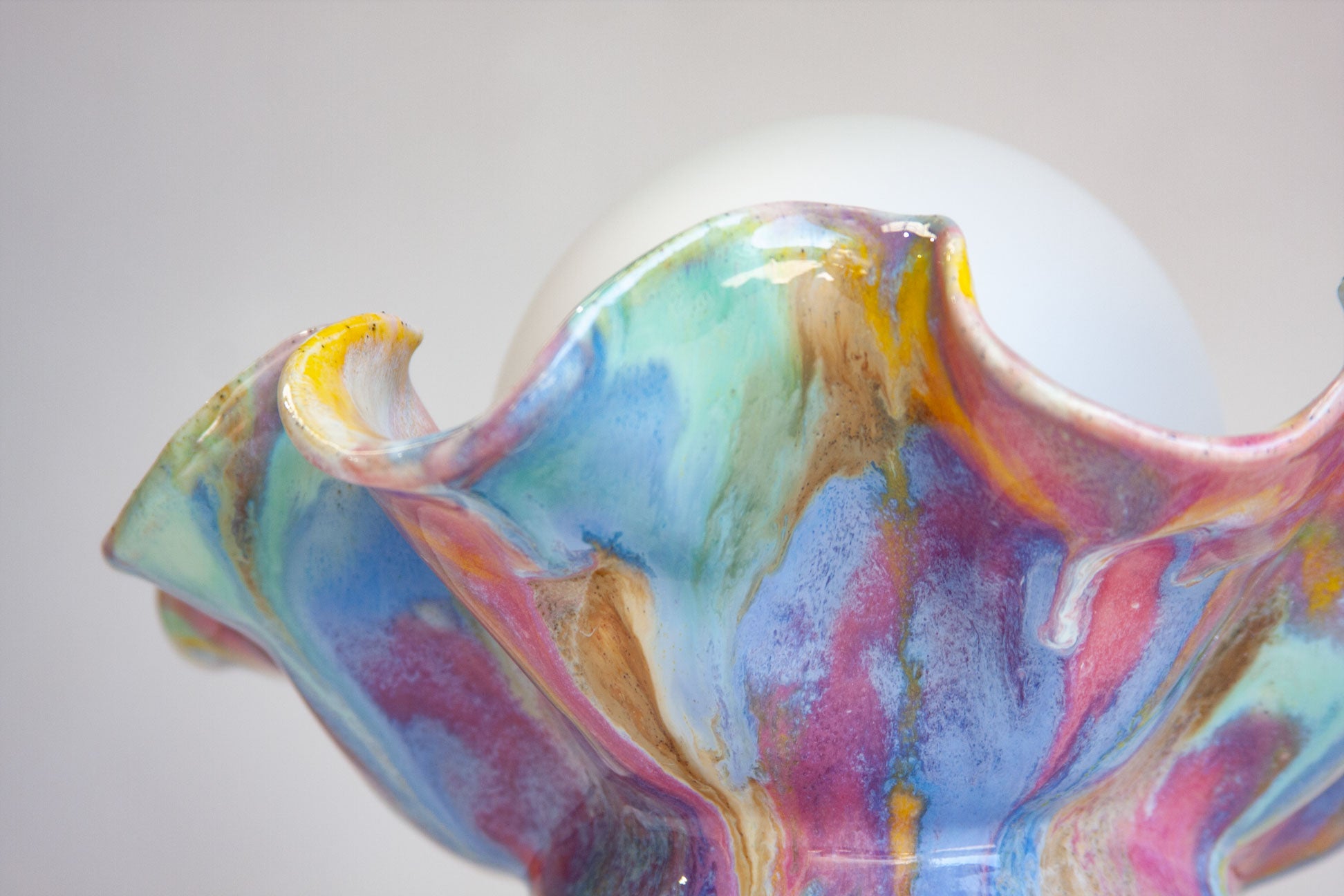A close-up of a colorful, ruffled decorative glass or ceramic bowl with marbled swirls of blue, pink, purple, green, and gold, with a white spherical insert visible behind it.