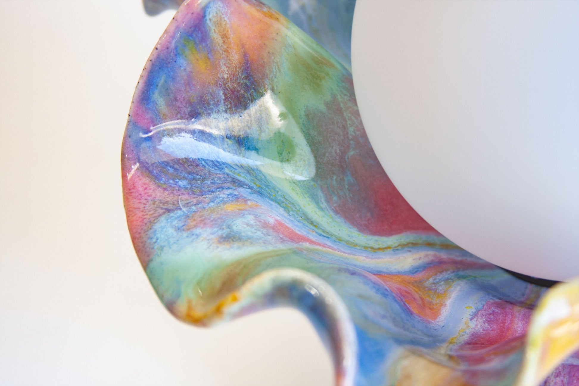 Close-up of a colorful marbled glass object with swirling blue, pink, green, purple, and orange tones, next to a smooth white curved surface.