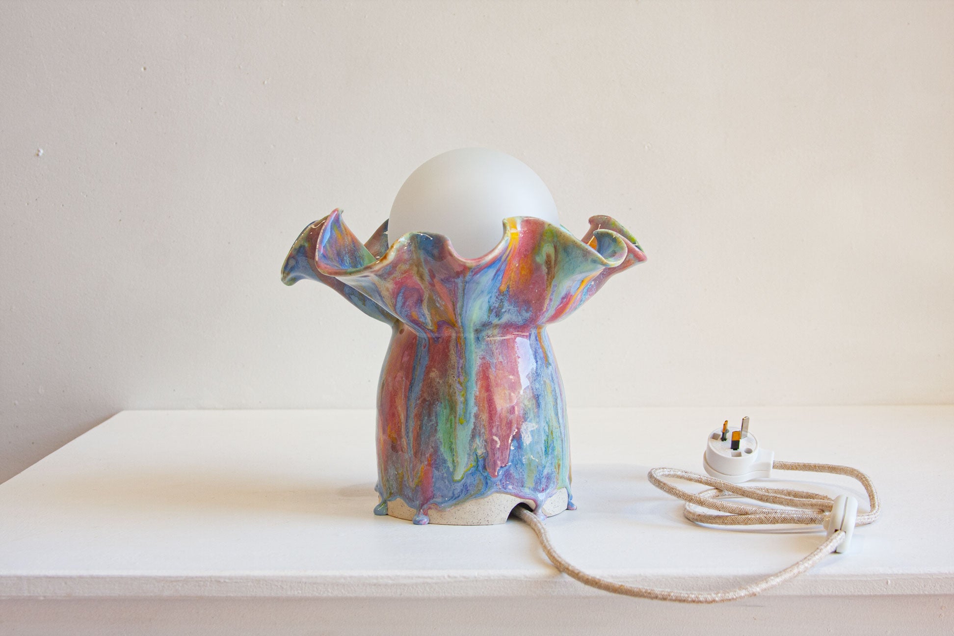 A handmade ceramic table lamp with a multicolored, ruffled body and a white glass globe shade, shown on a white shelf with its power cord and UK plug visible against a neutral beige background.
