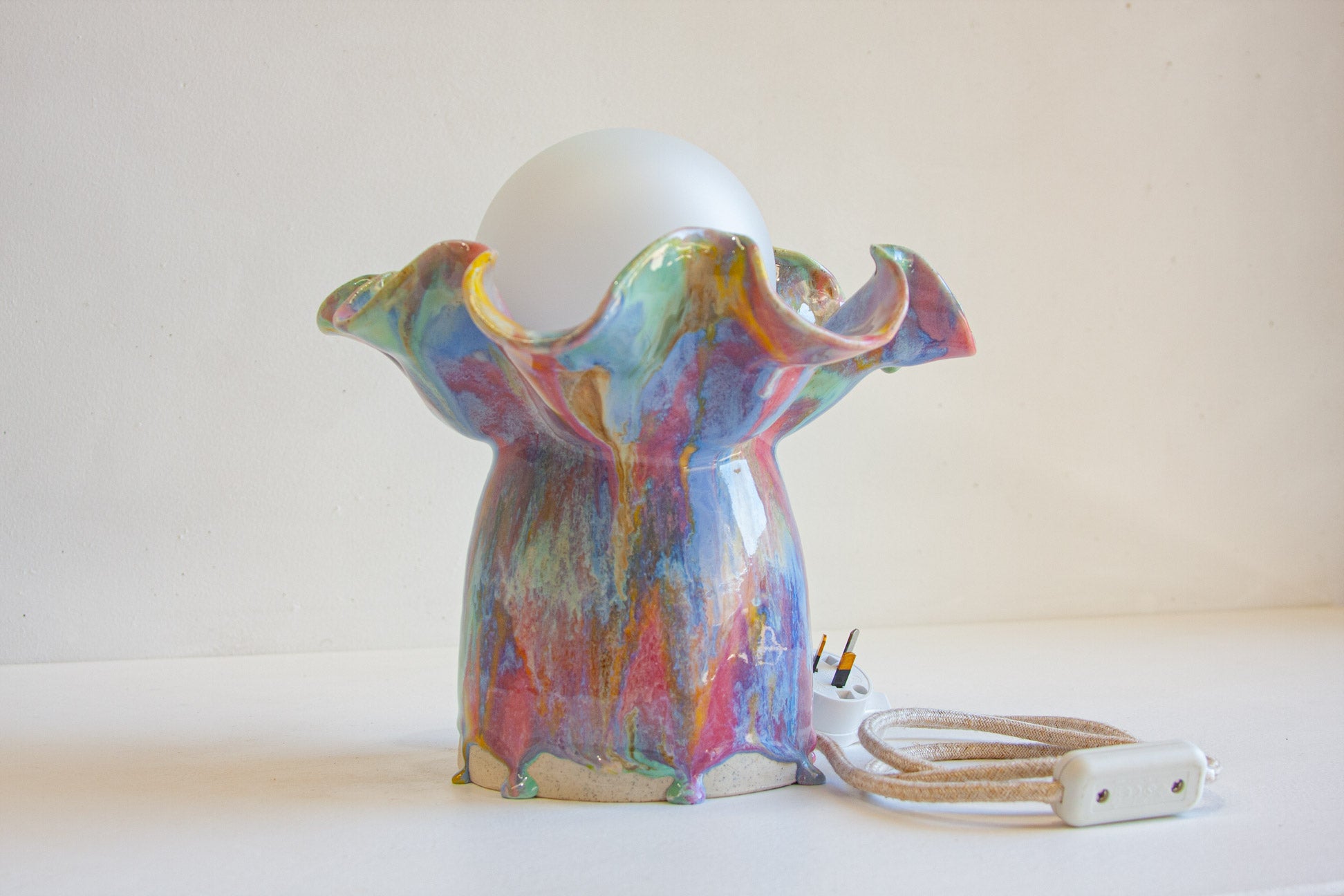 A colorful ceramic table lamp with a ruffled, flower-like base in a marbled multicolor glaze, holding a white globe shade. A beige cord with a white plug and switch extends from the base against a plain off-white background.