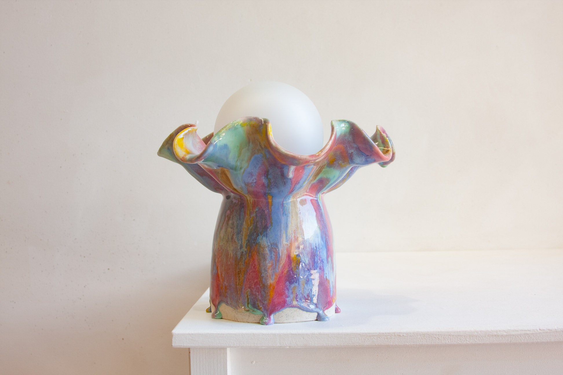 A colorful, sculptural ceramic table lamp base with a white spherical glass shade, displayed on a white shelf against a warm beige wall.