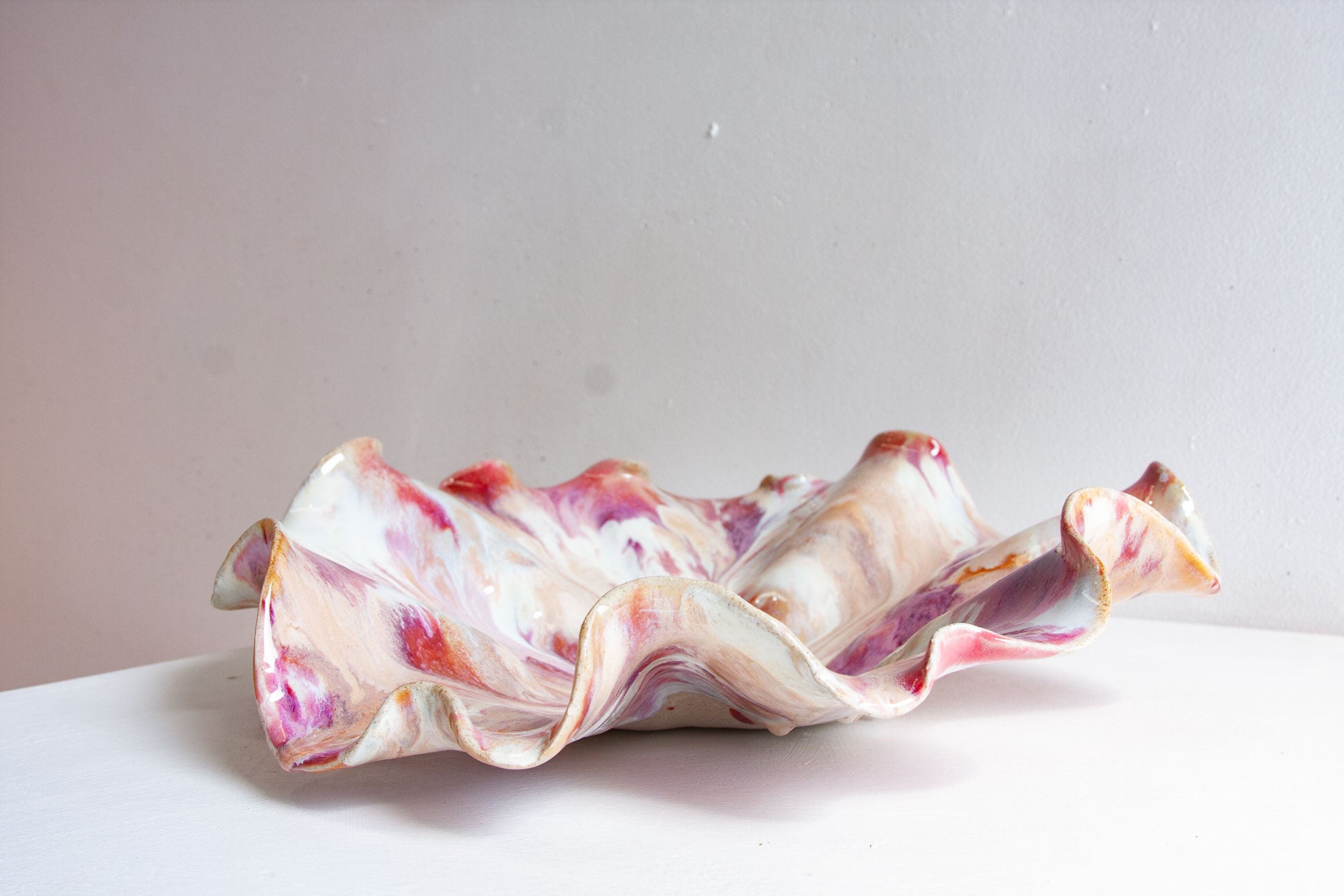 A handmade ceramic oyster platter seen from the side, its ruffled rim rising in waves, marbled in raspberry, plum, orange and cream.
