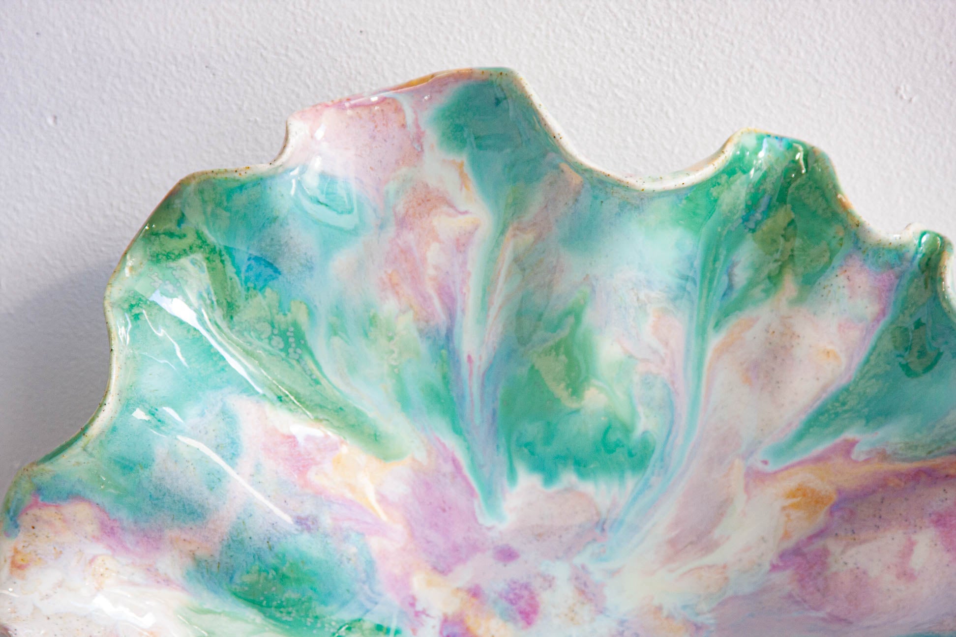 A close-up of a scalloped ceramic dish with a marbled, tie-dye-like glaze in pastel shades of green, pink, purple, and white against a plain light gray background.