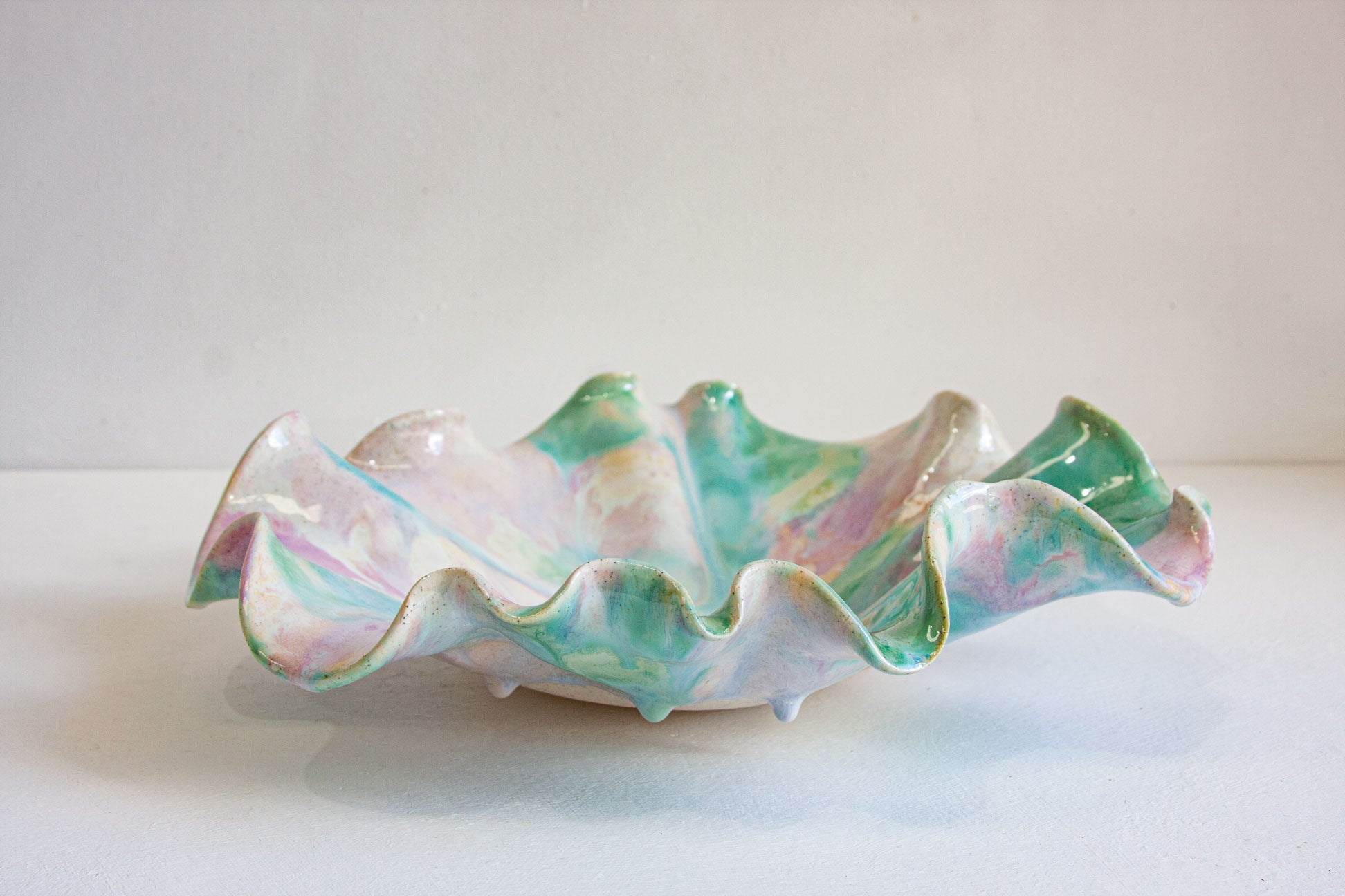 A handmade ceramic bowl with a wavy, ruffled edge and small feet, decorated with a marbled, iridescent glaze of pastel green, pink, blue, and white, set against a plain off-white background.