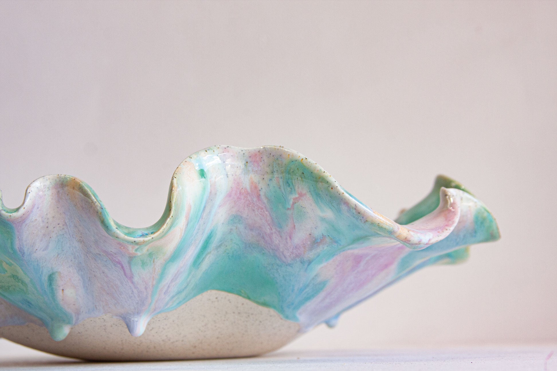 A close-up of a handmade ceramic bowl with a wavy rim, featuring a marbled pastel glaze of mint green, lavender, and pink over a speckled beige clay base.