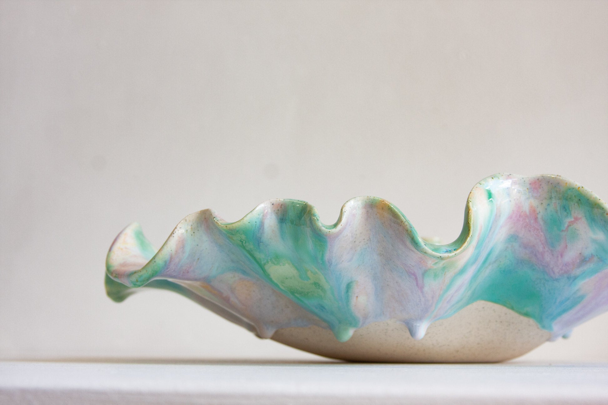 A handmade ceramic bowl with a wavy, scalloped rim, featuring a marbled glaze of pastel turquoise, green, lavender, and pink over a warm beige base, set against a neutral light gray background.