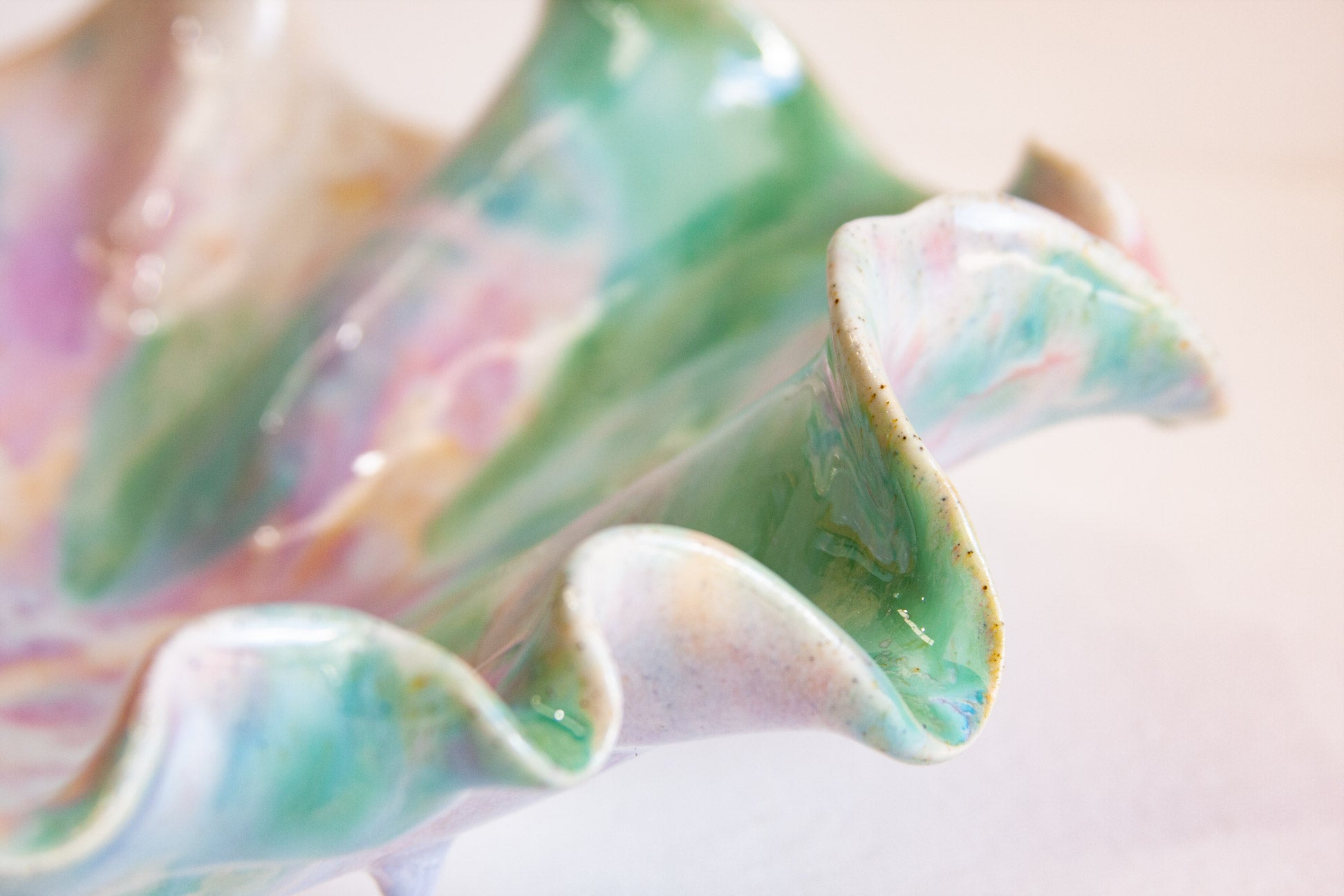 A close-up of a glossy ceramic shell-shaped vessel with wavy ruffled edges, featuring marbled pastel green, pink, and cream glaze on a soft light beige background.