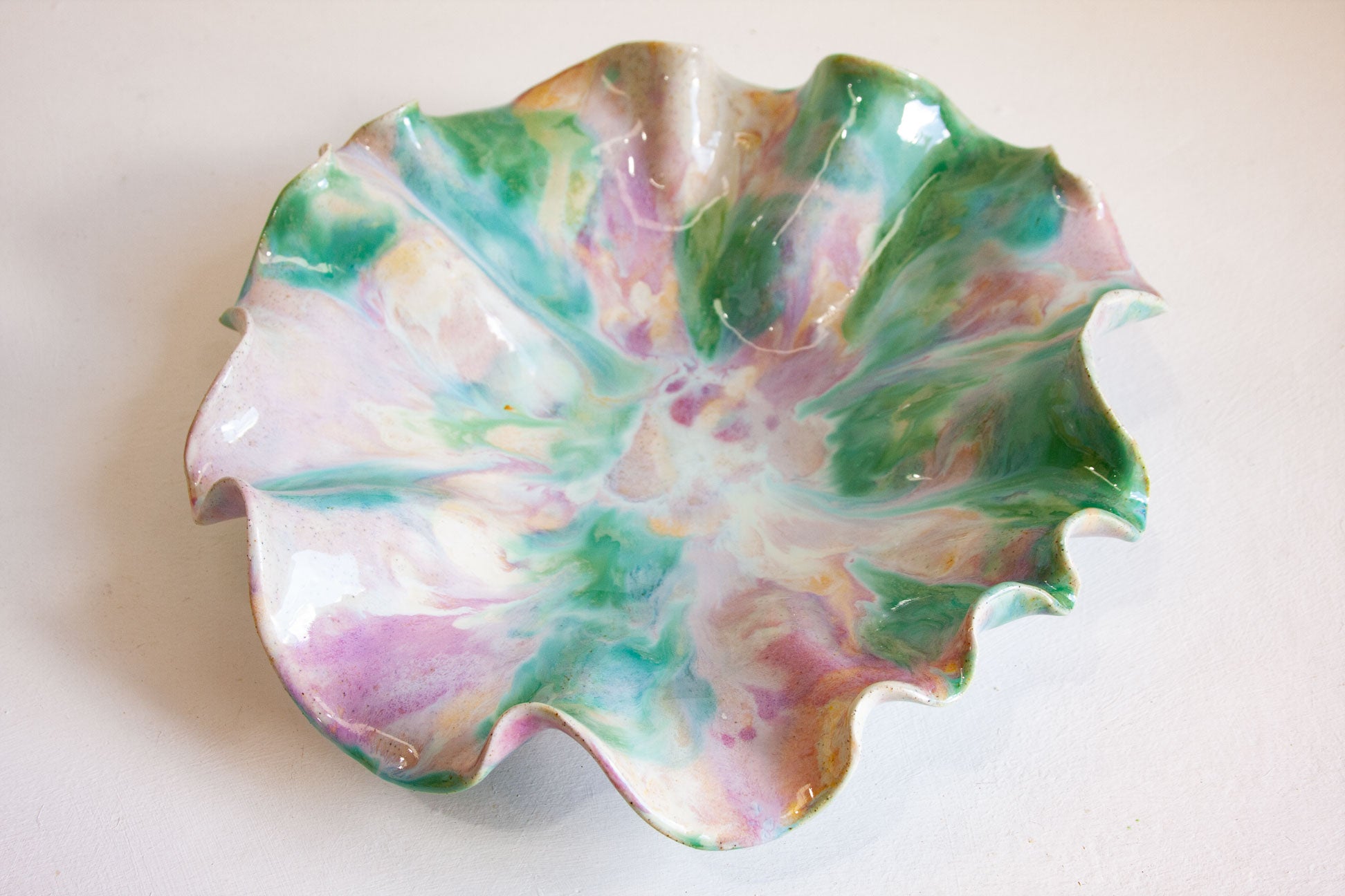 A handmade ceramic bowl with a wavy, petal-like rim, decorated with a marbled pattern of green, pink, purple, and white glaze on a light beige background.