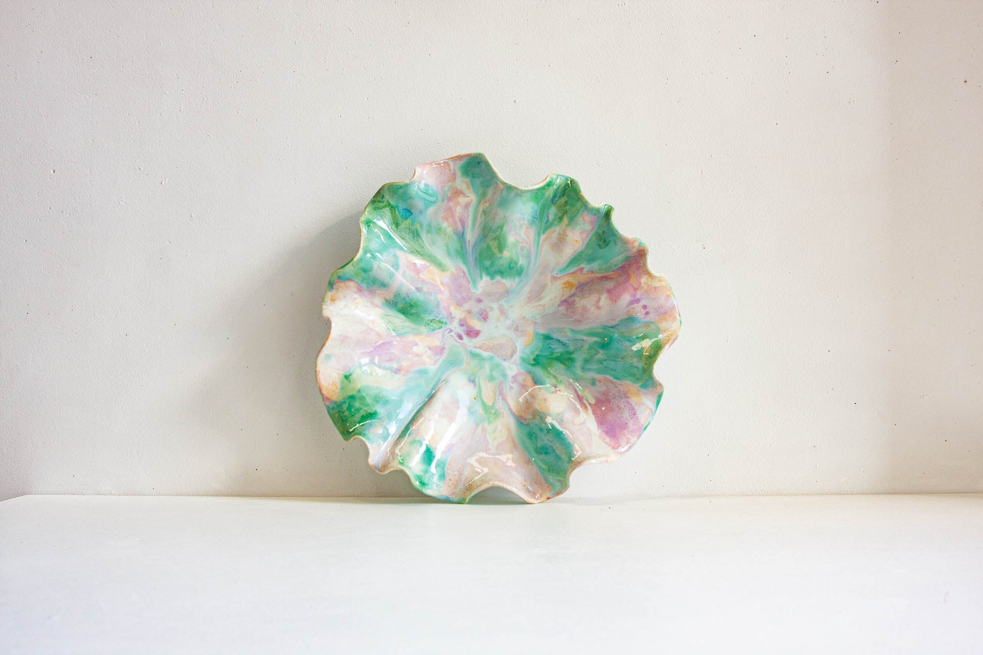 A scalloped ceramic plate with a marbled pattern of green, pink, and white, displayed against a plain off-white background.