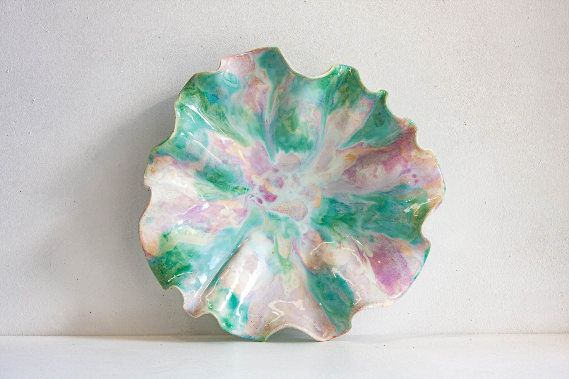 A scalloped ceramic plate with a marbled floral pattern in shades of green, pink, and white, set against a plain off-white background.