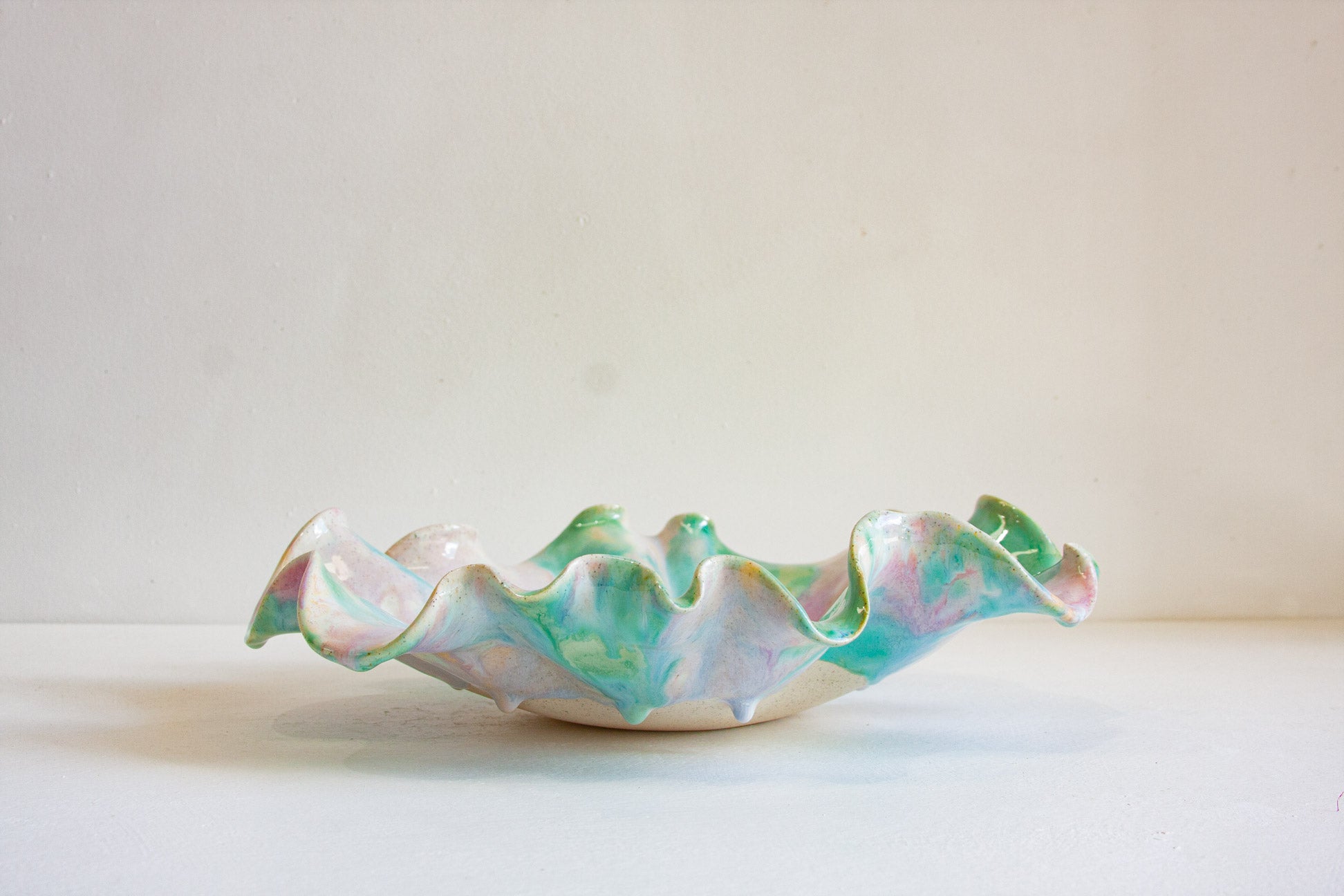 A handmade ceramic bowl with a ruffled, scalloped edge, featuring a marbled glaze of pastel green, pink, and white on a cream base, set against a plain off-white background.