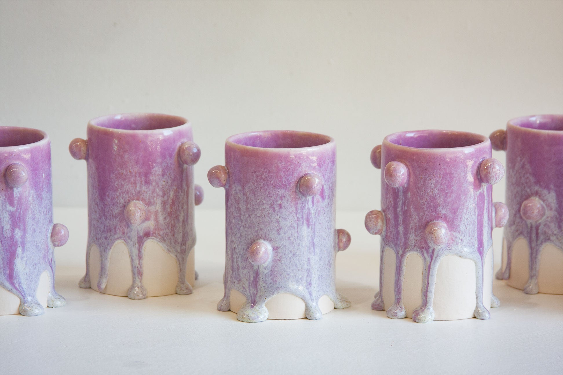 A row of small ceramic cups with a purple and white drip glaze design, featuring raised circular details on the sides, set against a plain off-white background.