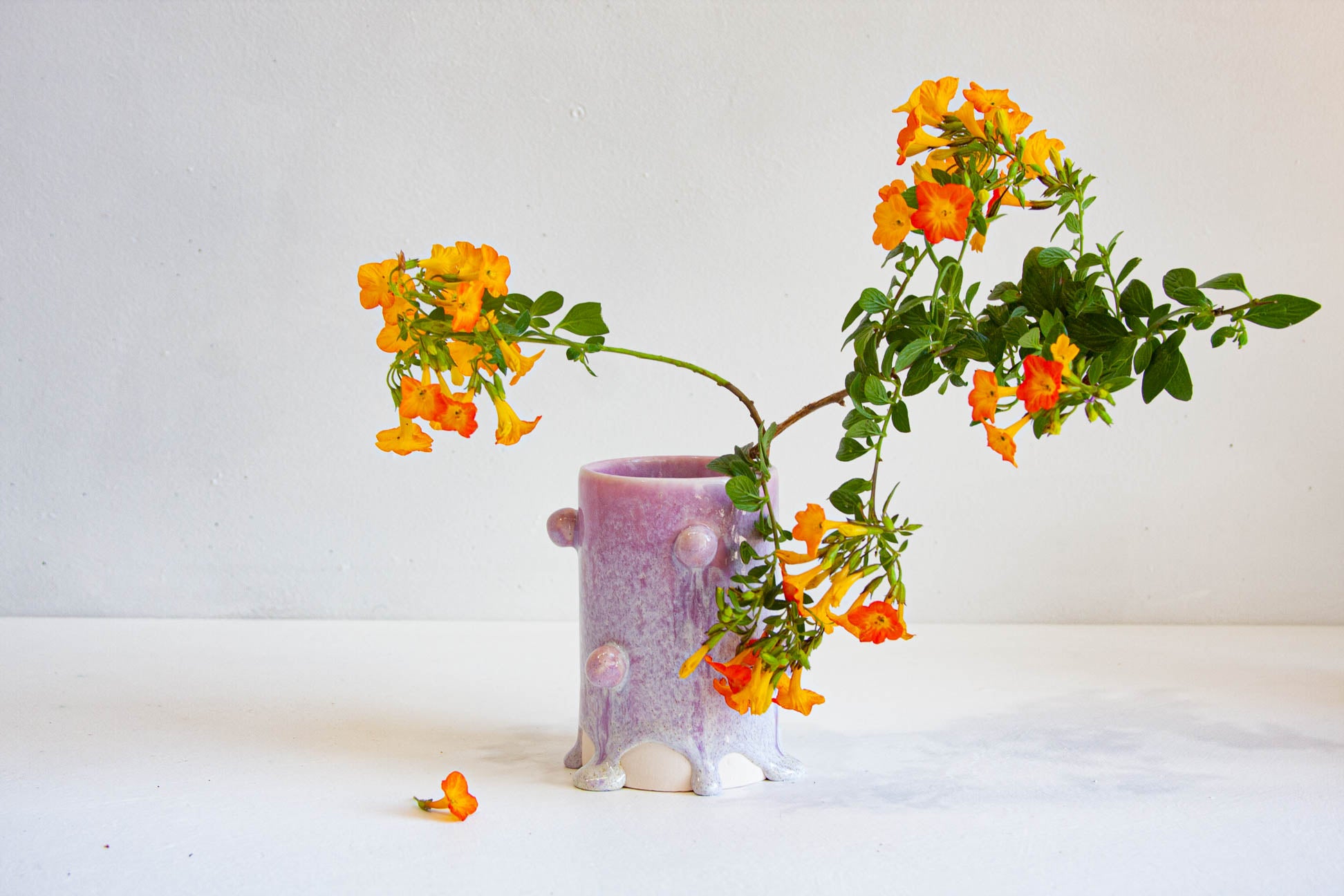 A small lavender ceramic pot with a textured, bubbly surface and tiny raised feet, holding a flowering branch with bright orange blossoms and green leaves, set against a plain light background.