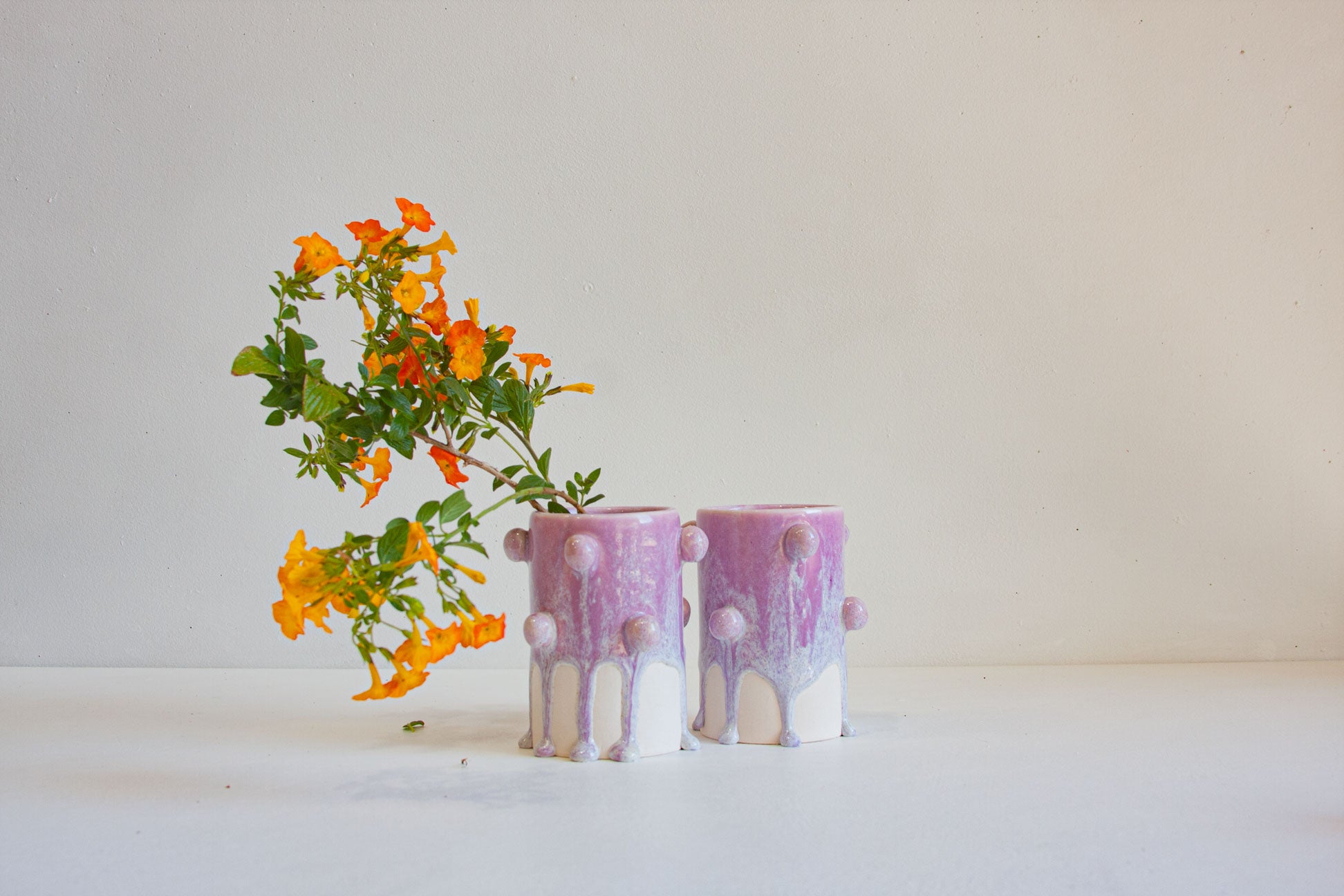 Two small cylindrical ceramic vases with a purple and white drip glaze finish, holding bright orange flowers, set against a plain off-white background.