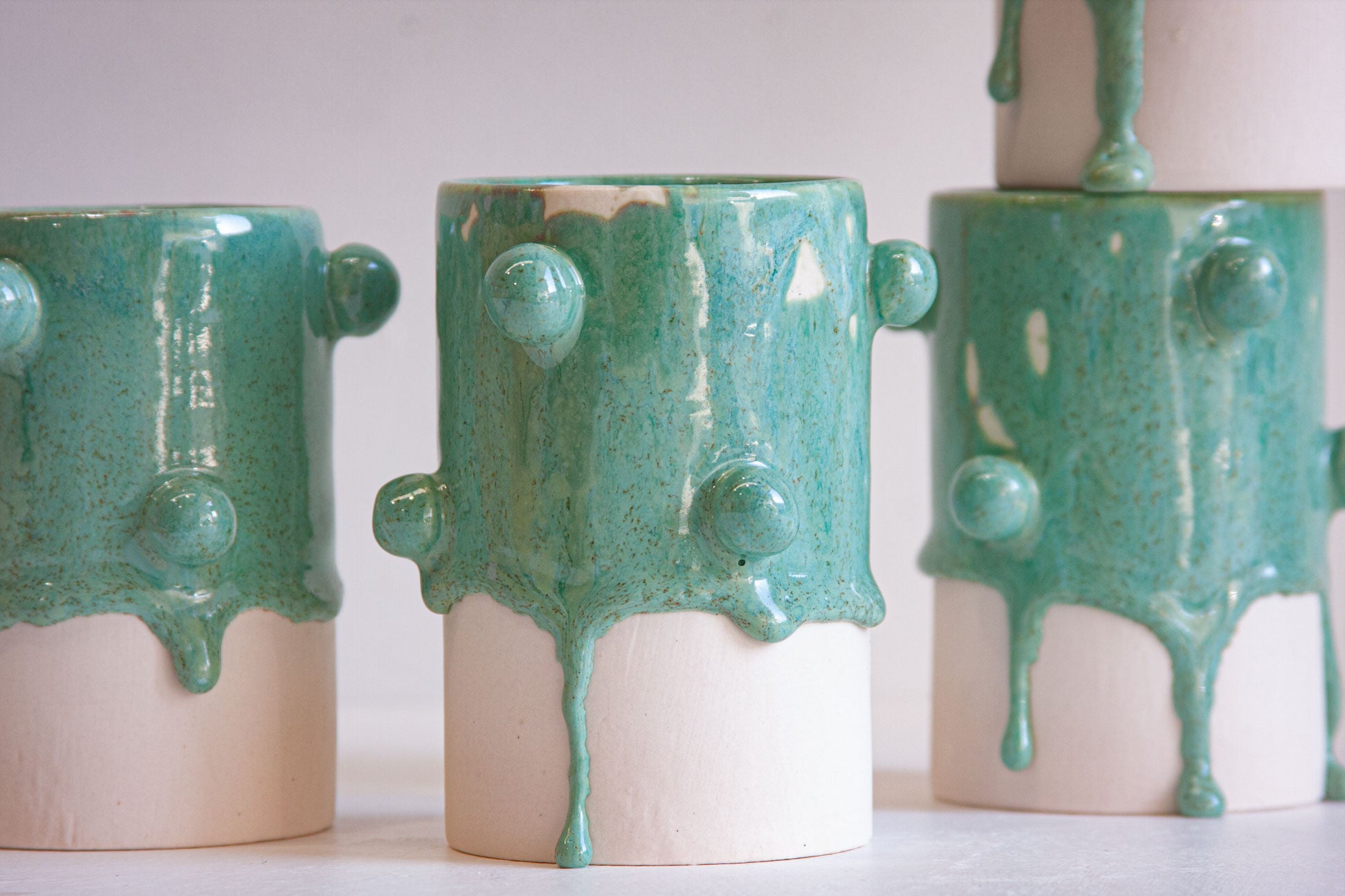 Three handmade ceramic vases with a glossy teal-green dripping glaze and a matte off-white base, arranged on a light surface against a pale background.