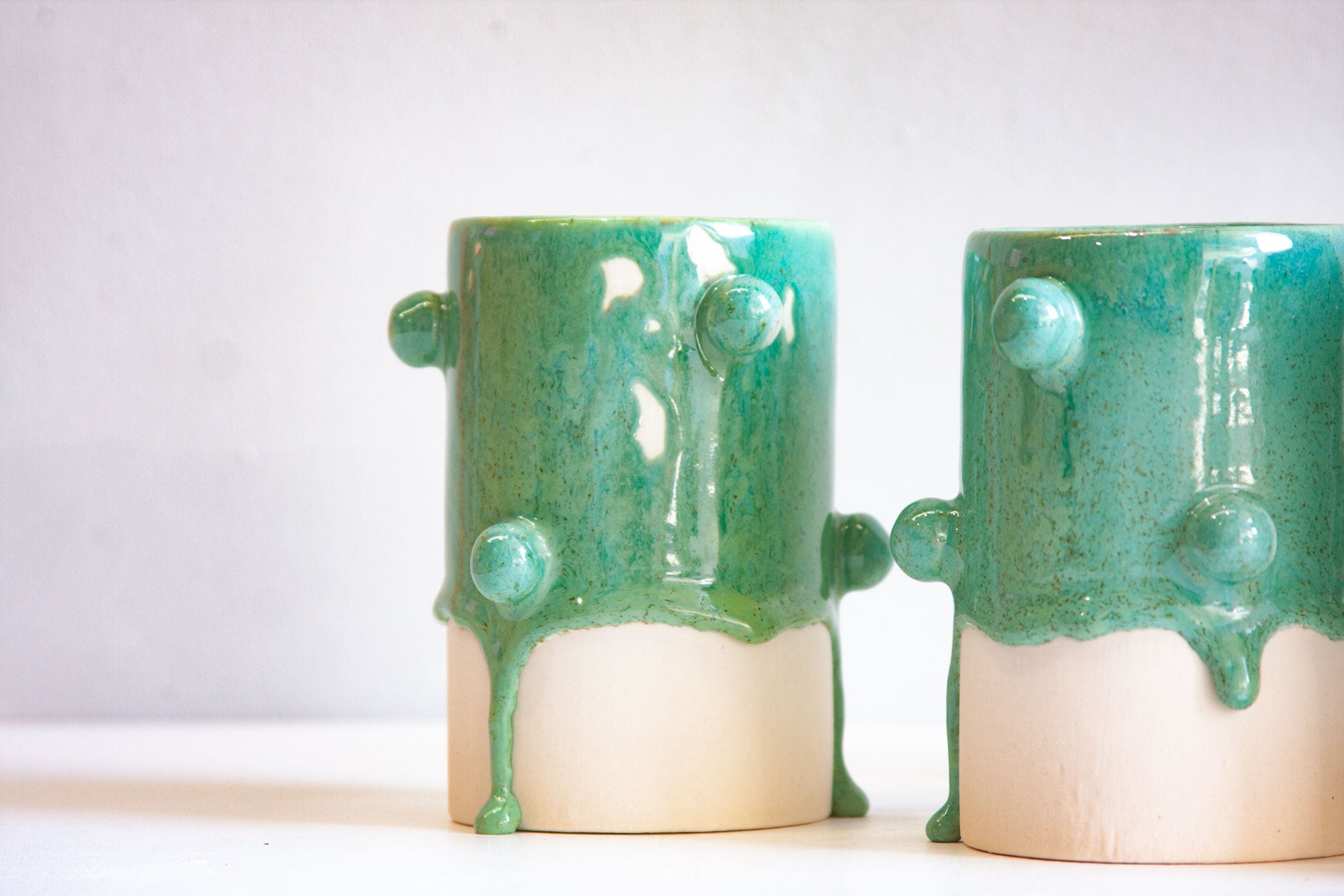 Two handmade ceramic cups with a glossy dripping green glaze and raised circular details, shown on a plain light background.