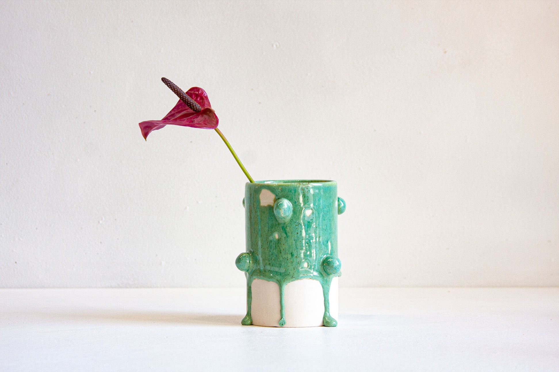 A small cylindrical ceramic vase with a glossy green and white drip design, holding a single magenta anthurium flower, set against a plain off-white background.