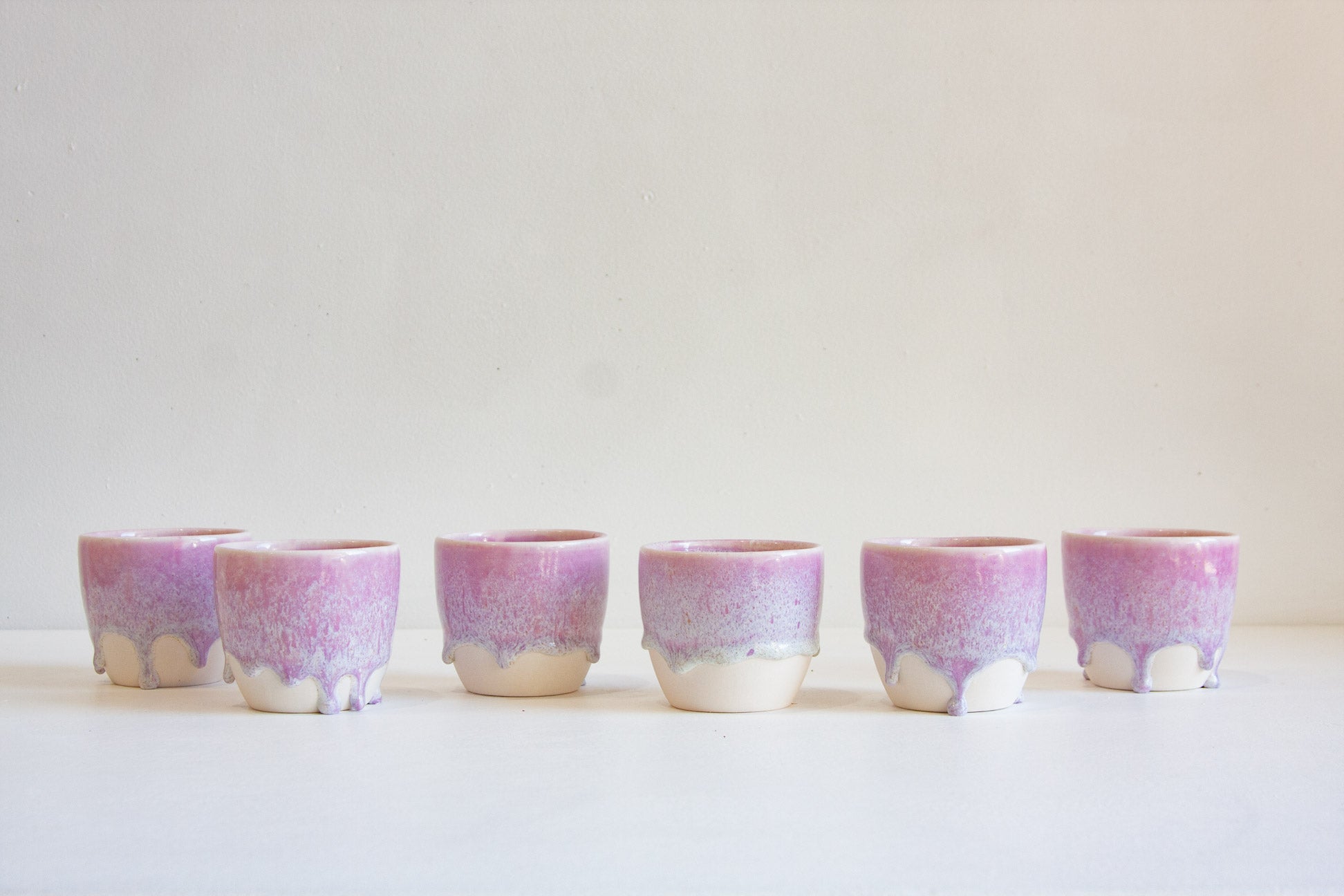 Six small ceramic cups with a purple and pink gradient glaze and dripping white base, arranged in a row against a plain light background.