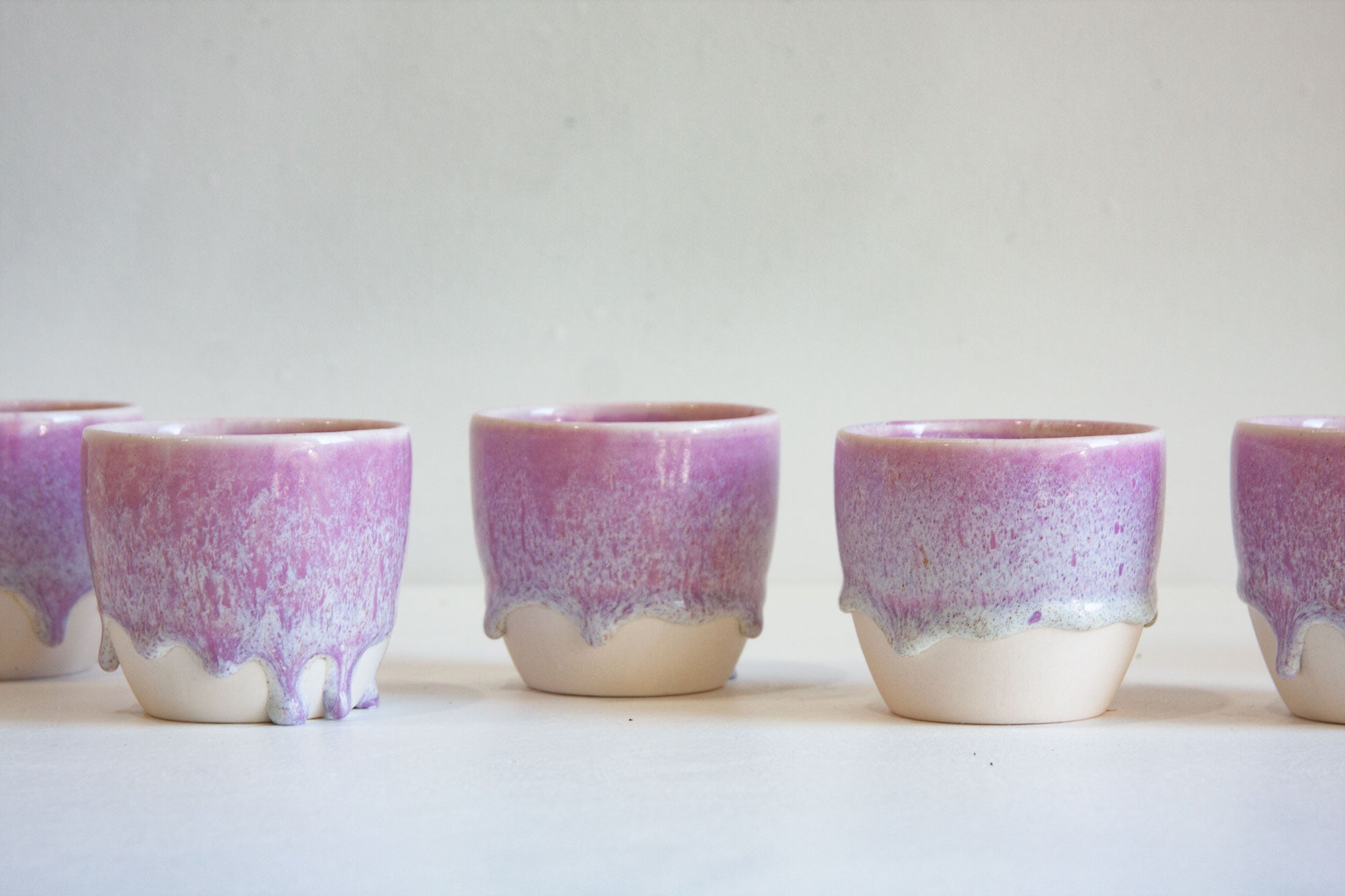 A row of small ceramic cups with a purple and pink glaze dripping over a cream-colored base, set against a plain light background.