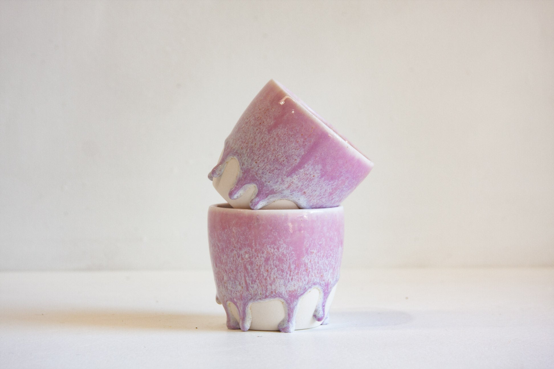 Two small ceramic cups stacked with one tilted on top of the other, featuring a pink and white dripping glaze pattern, set against a plain off-white background.