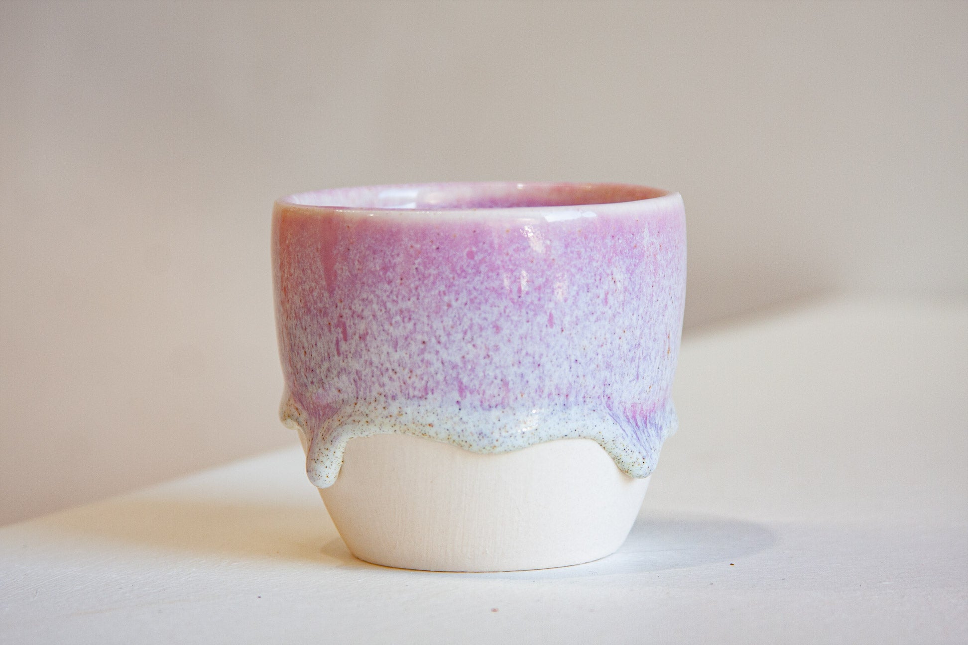 A handmade ceramic cup with a pink and purple speckled glaze on the upper half and a dripping white base, set against a neutral beige background.