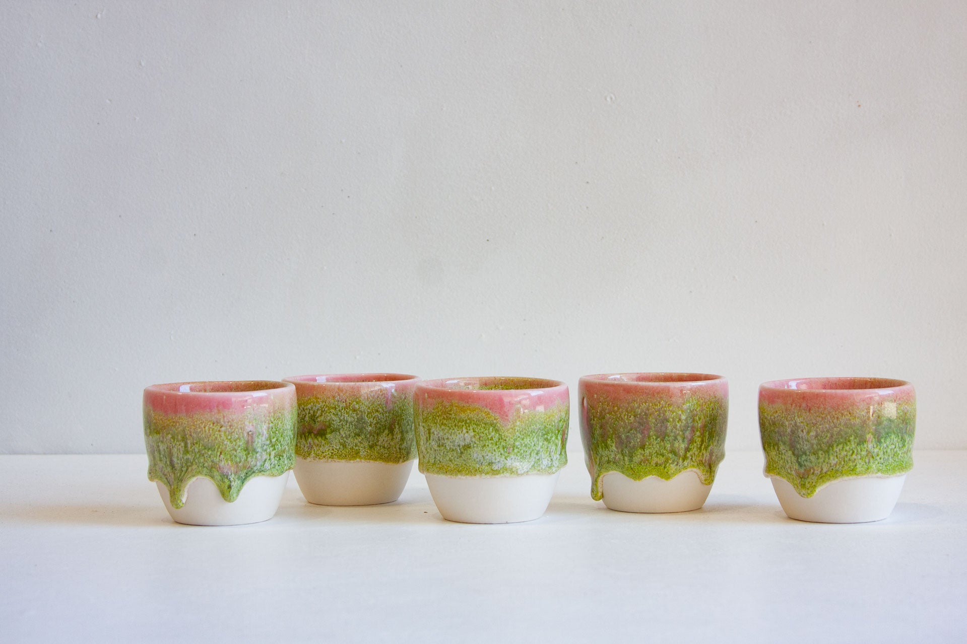 Six small ceramic cups arranged in a row on a white surface against a light gray background. Each cup has a glossy dripping glaze pattern with pink at the top, green in the middle, and a white base.