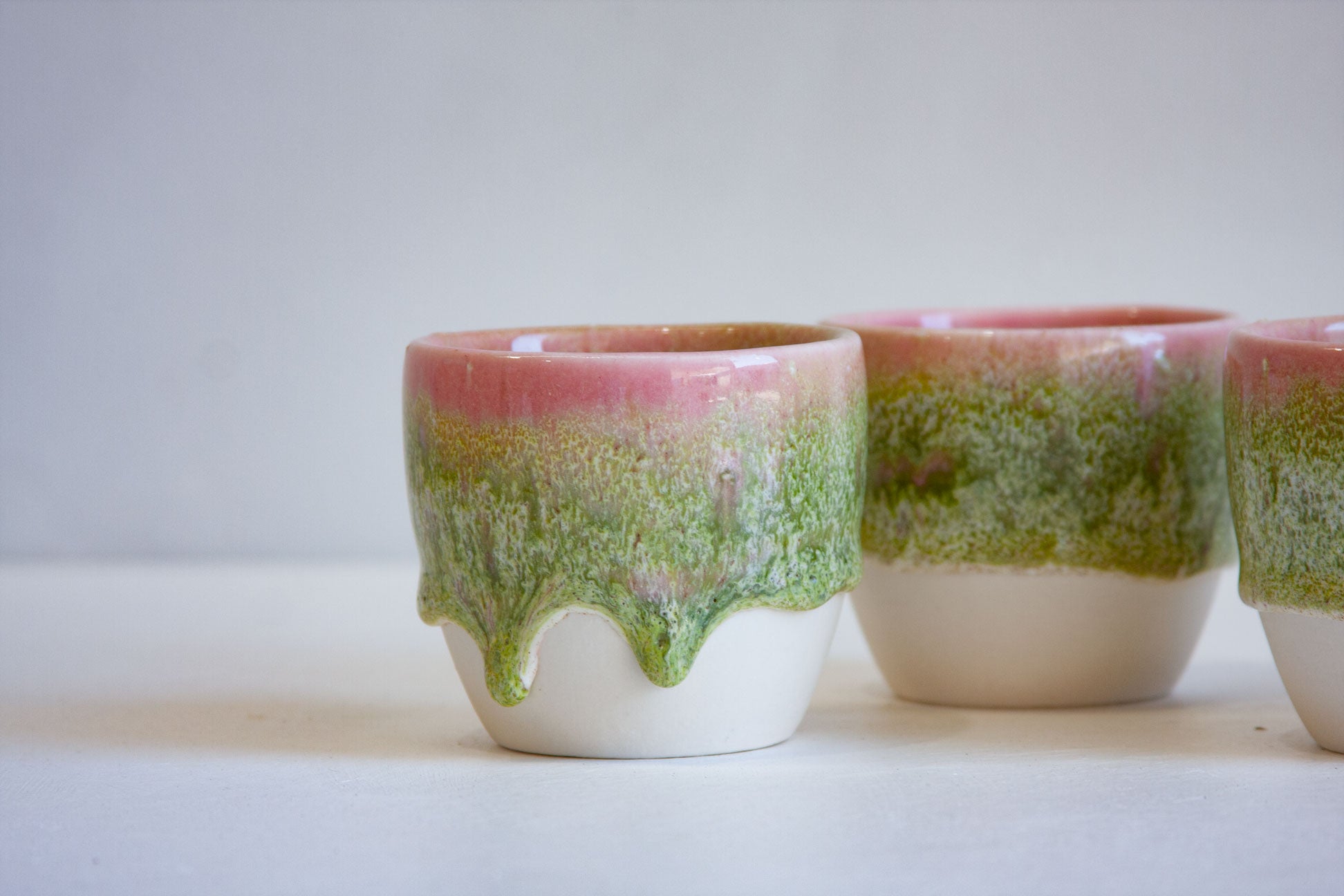Three small ceramic cups with a dripping glaze effect, featuring a pink top section, a textured green middle section, and a white bottom, set against a plain light gray background.