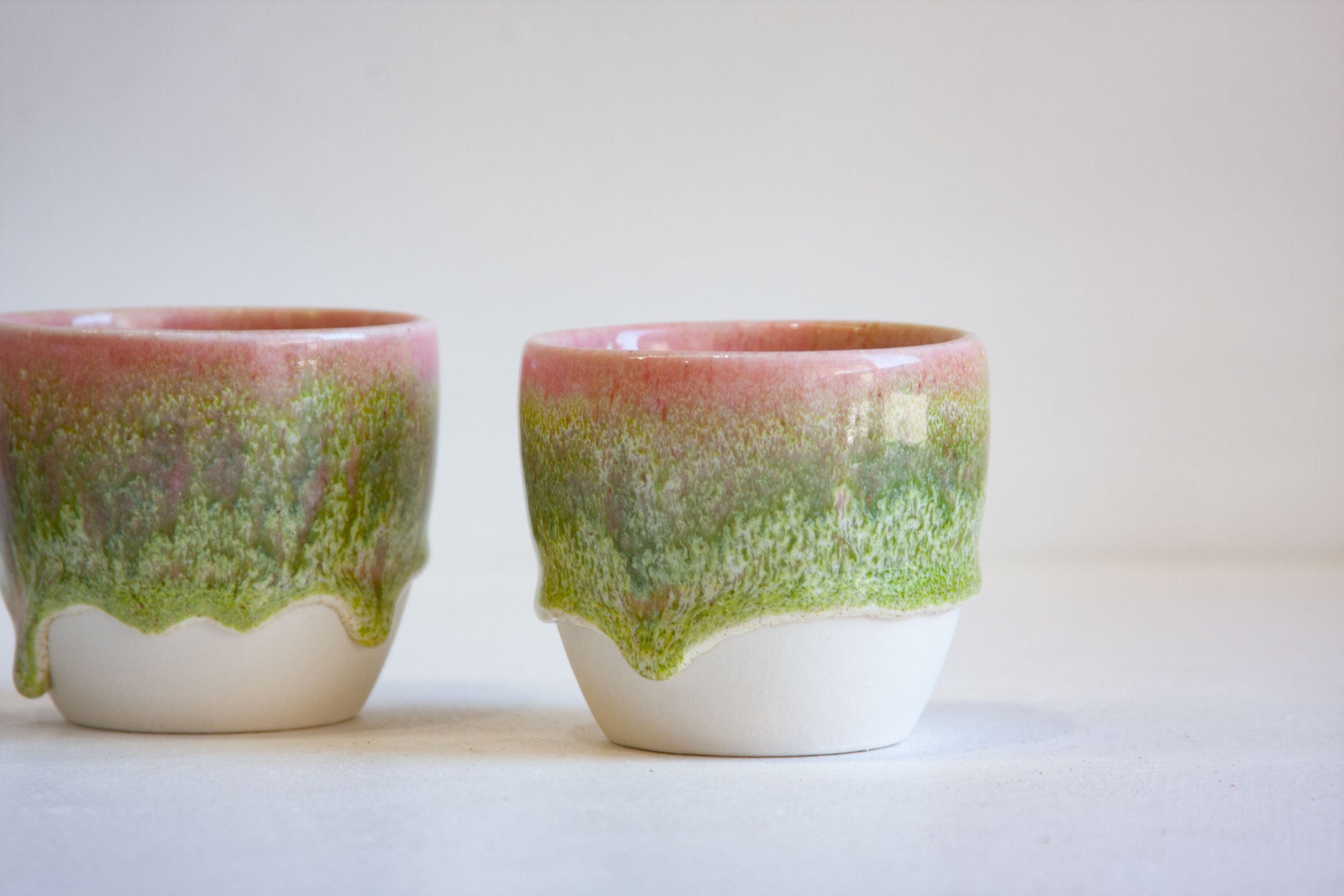 Two handmade ceramic cups with a dripping glaze design, featuring a pink top section, a green middle section, and a white bottom, set against a plain light background.