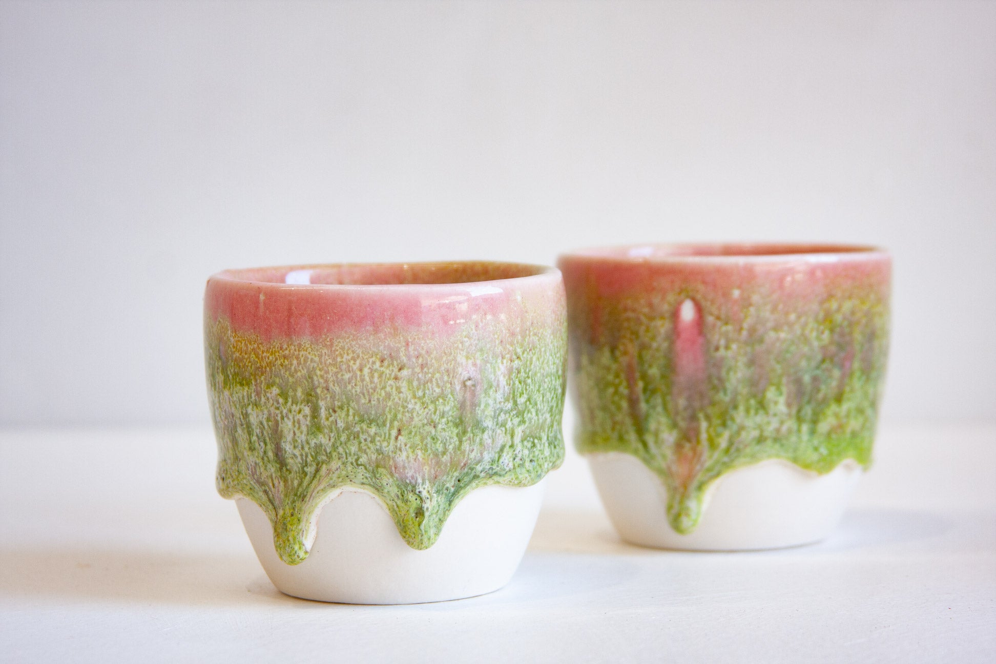 Two small ceramic cups with a dripping glaze design, featuring a pink top section, a green middle section, and a white bottom, set against a plain light background.