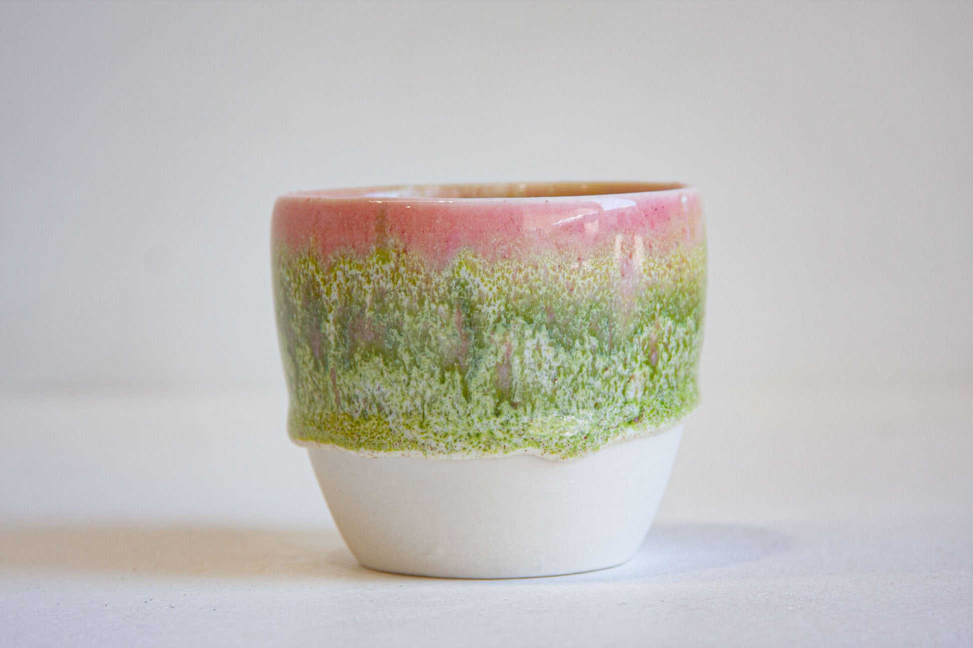 A handmade ceramic cup with a pink and green glazed upper half and a solid white unglazed lower half, set against a plain light gray background.