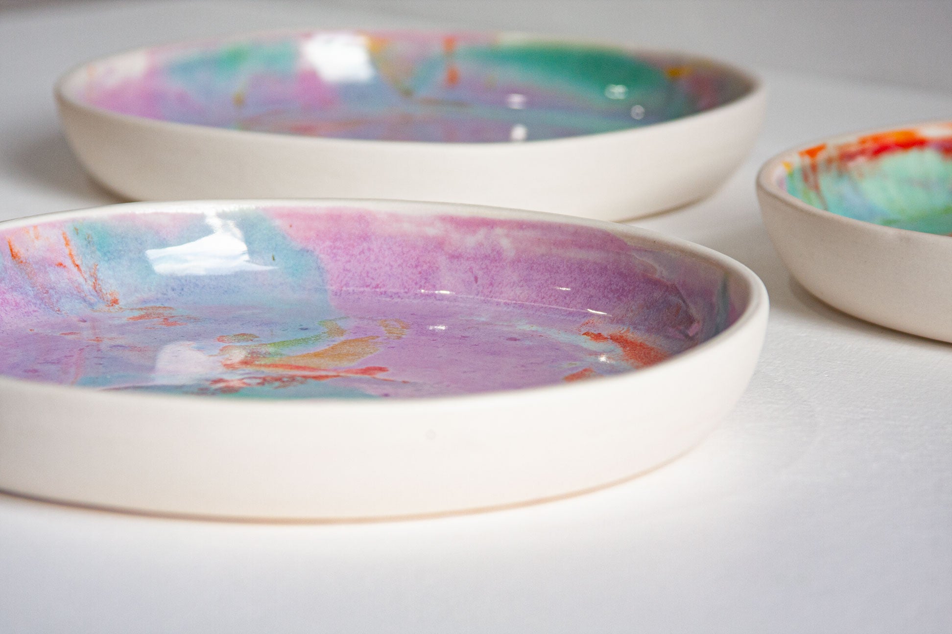 Three shallow ceramic bowls with a cream-coloured exterior and colourful marbled interiors in pink, purple, blue, green, and orange, arranged on a white surface.