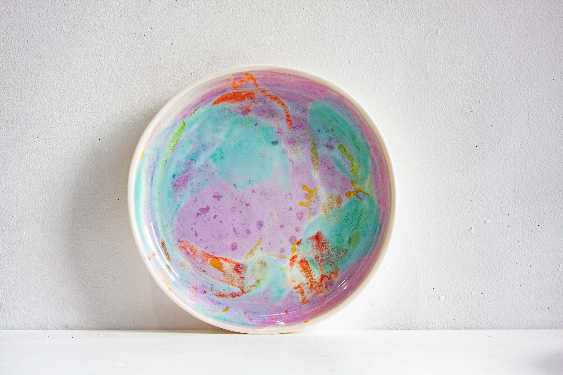 A round ceramic plate with a colourful marbled pattern in pastel purple, teal, and orange, set against a plain white wall.