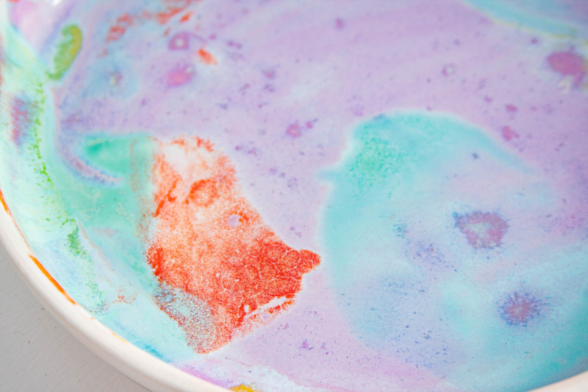 A close-up of a colorful, marbled surface with swirling pastel purple, light blue, mint green, and orange-red areas, partially framed by a white curved edge.
