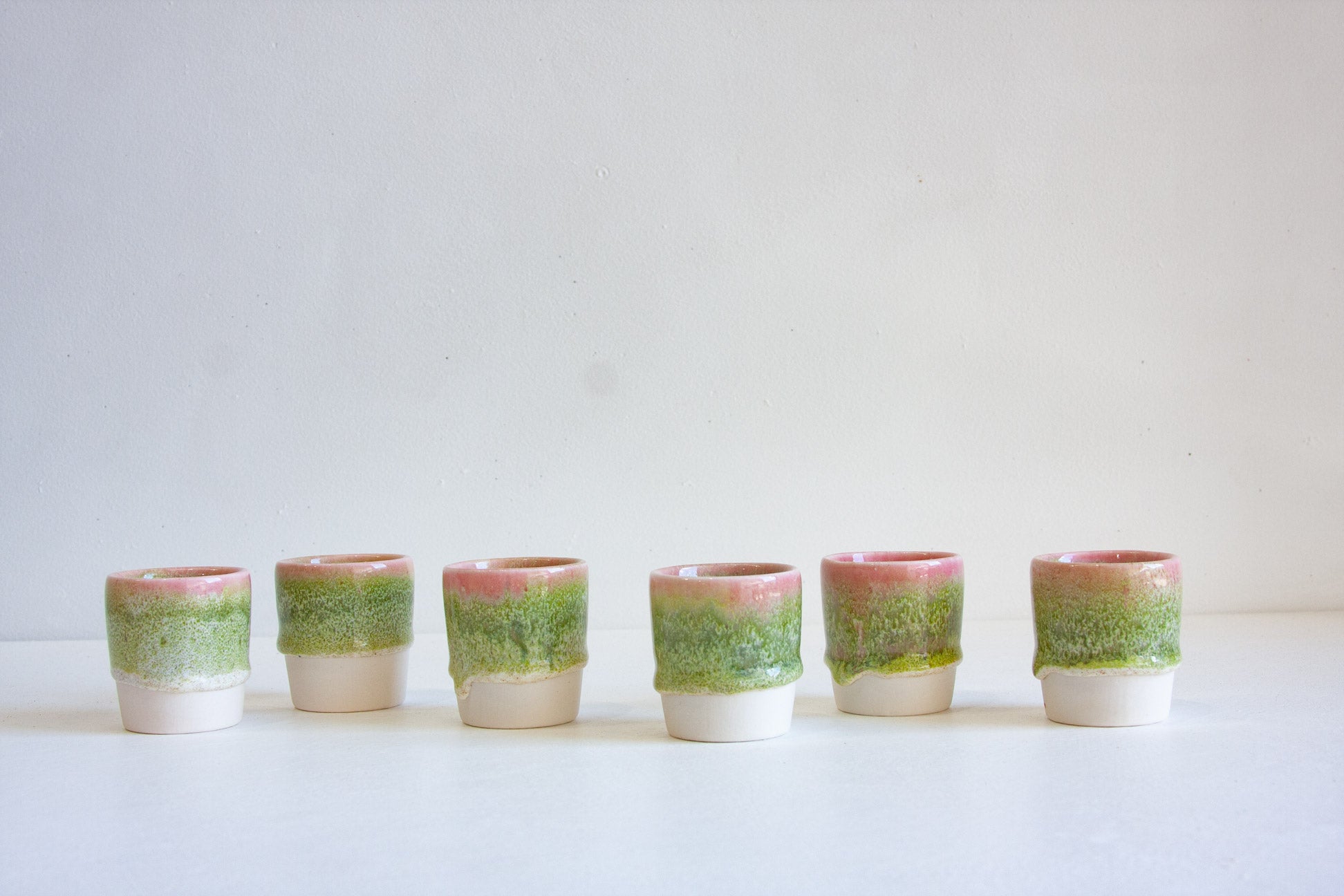 Six small ceramic cups lined up in a row on a white surface against a light gray background. Each cup has a textured green and pink glazed upper half and a plain white lower half.