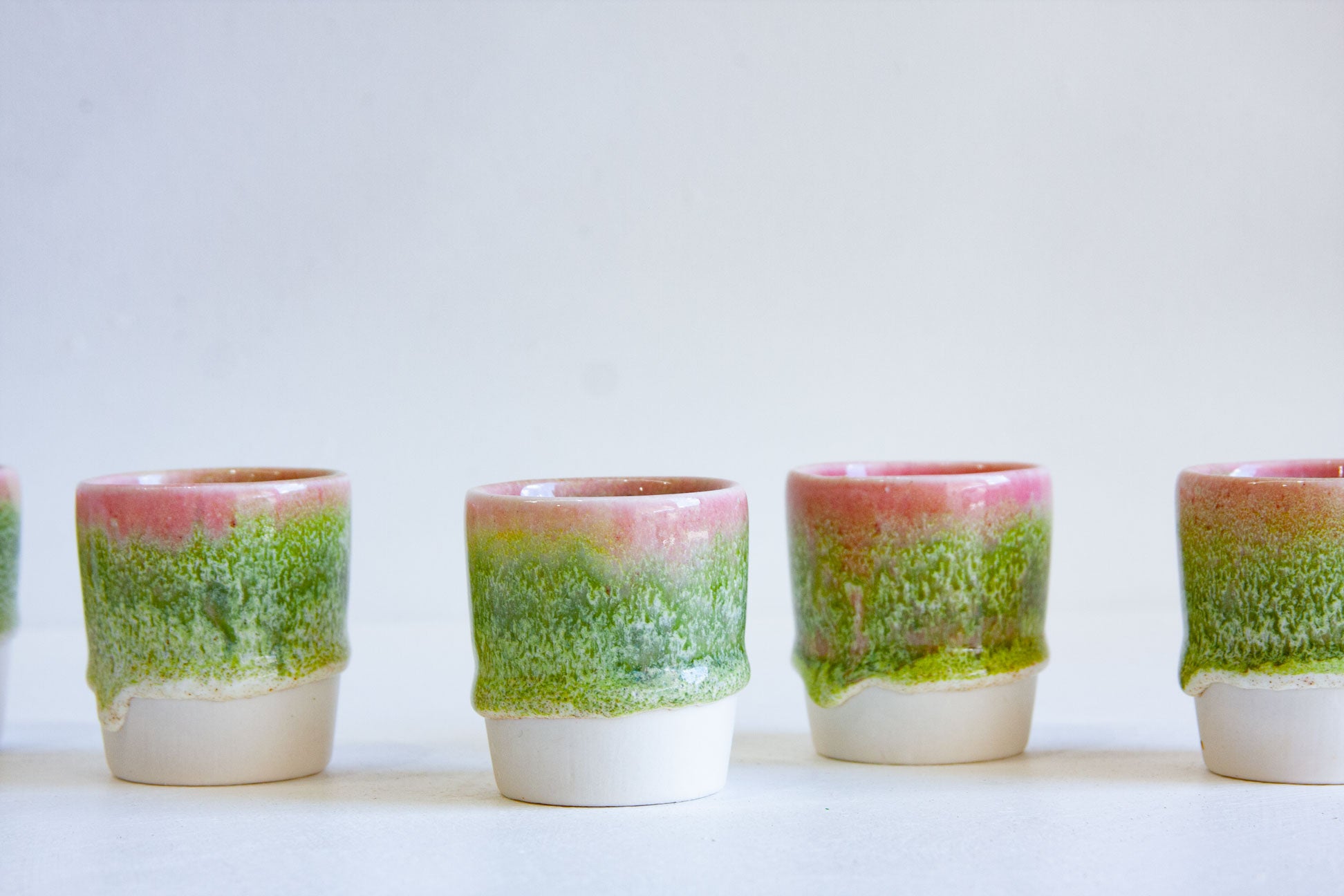A row of small ceramic tumblers with a dripping glaze effect, featuring a vibrant green and pink upper section and a matte white lower base, set against a plain white background.