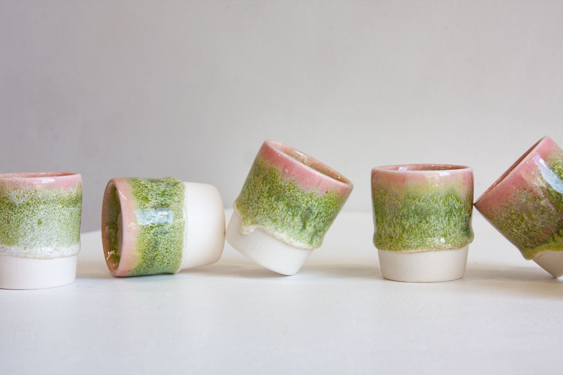 Five handmade ceramic tumblers with a green and pink speckled glaze and a solid white base, arranged in a row on a light gray surface.