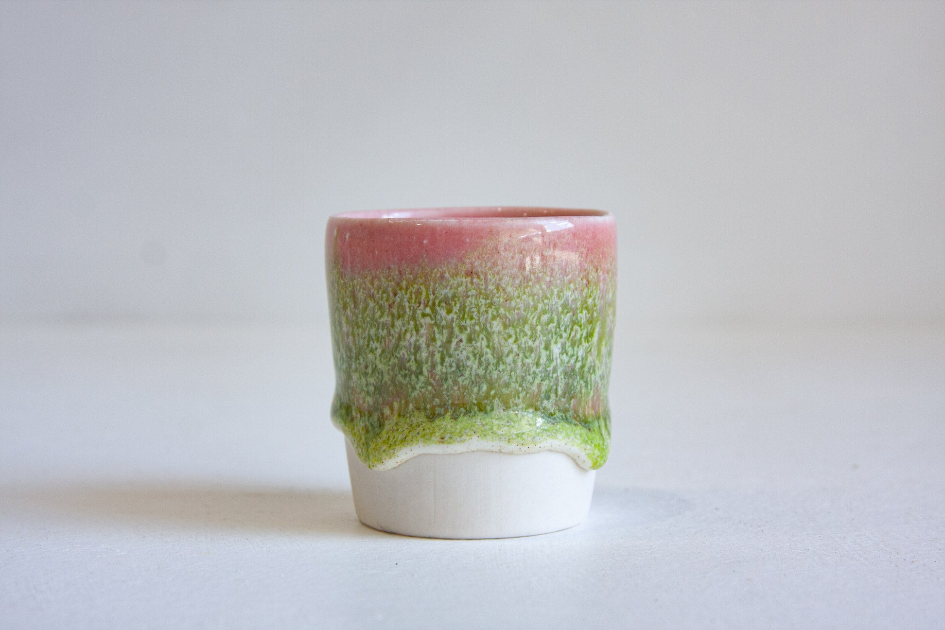 A handmade ceramic cup with a dripping glaze design, featuring a pink top section, a textured green middle section, and a white base, set against a plain light gray background.