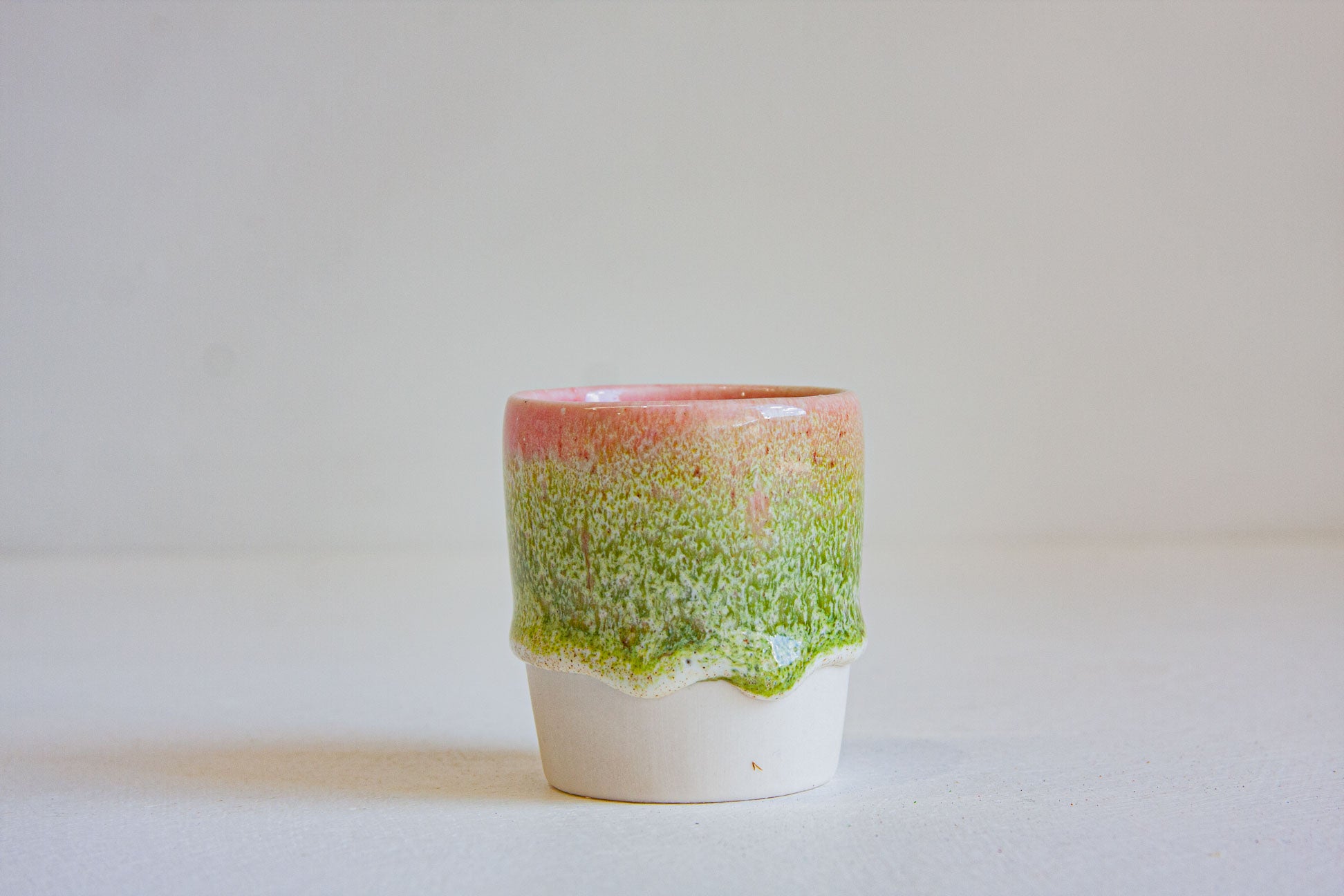 A small handmade ceramic cup with a dripping glaze design, featuring a pink top section, a green speckled middle section, and a white base, set against a plain light background.