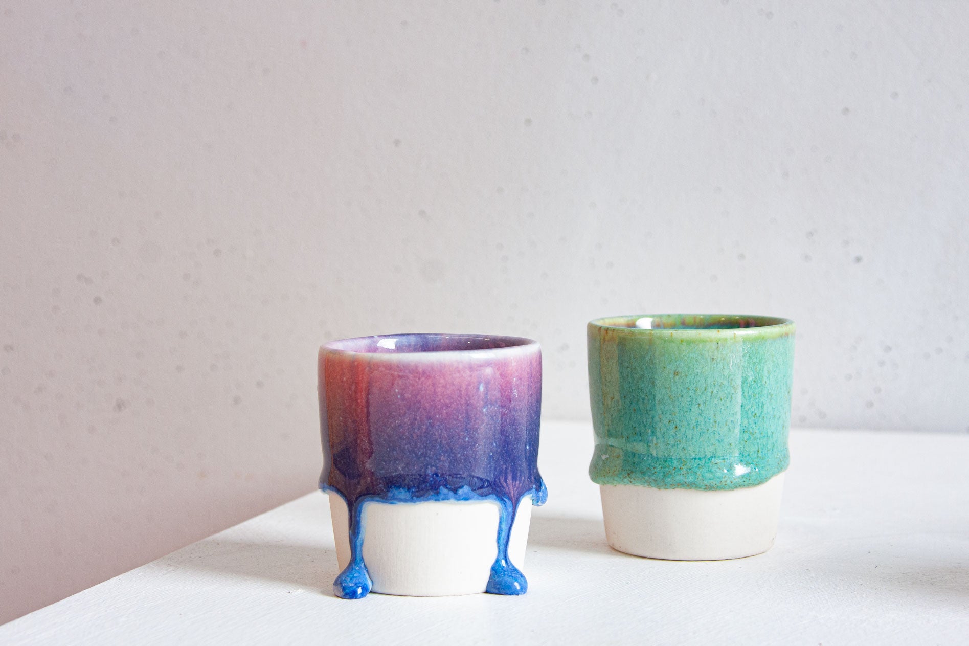 Two handmade ceramic cups, one glazed in purple and magenta fading into blue with bright drips at the foot, the other a jade green with a yellow-green rim.