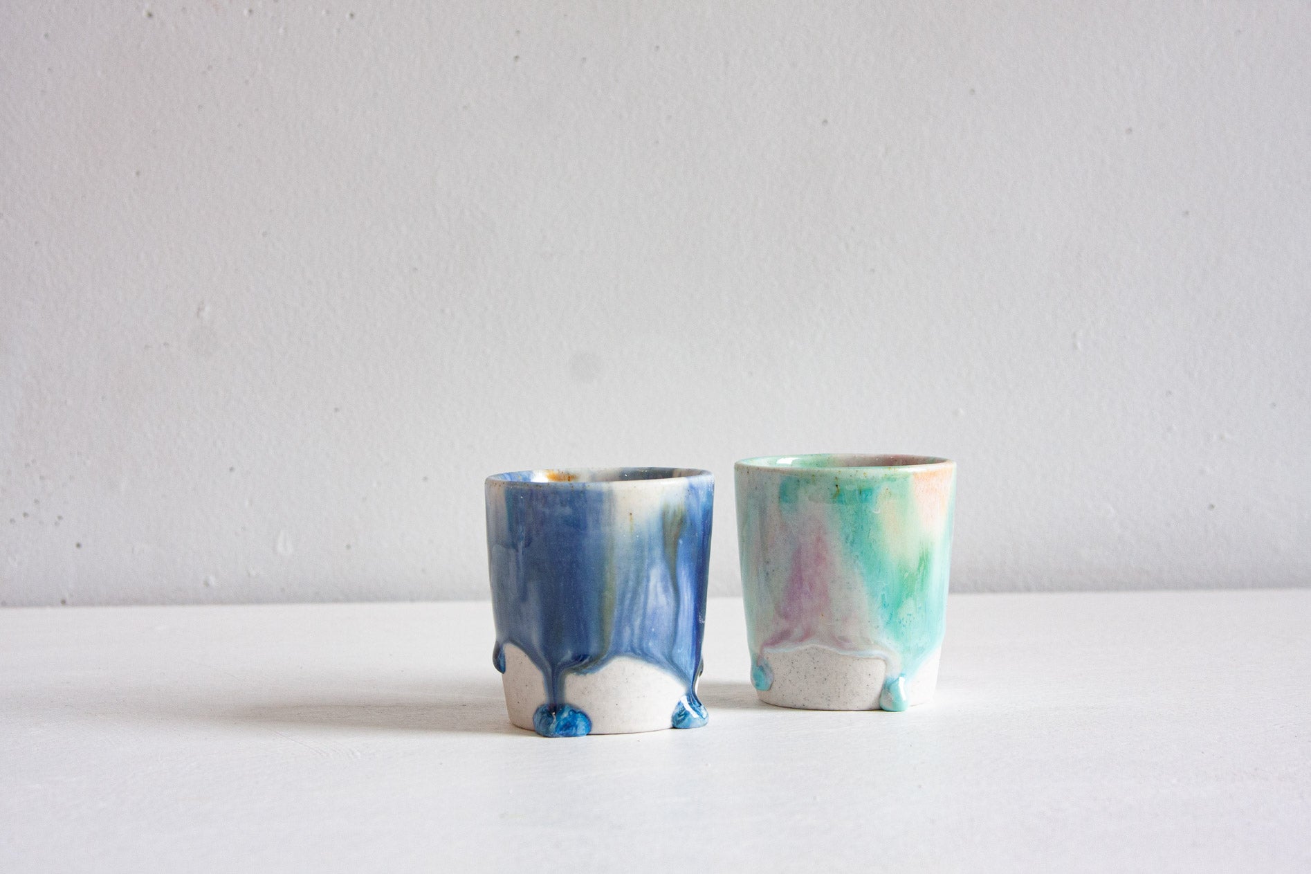 Two handmade ceramic cups side by side, one in deep blue running through ochre and white, the other a soft mint flushed with pink and peach, over bare clay bases.