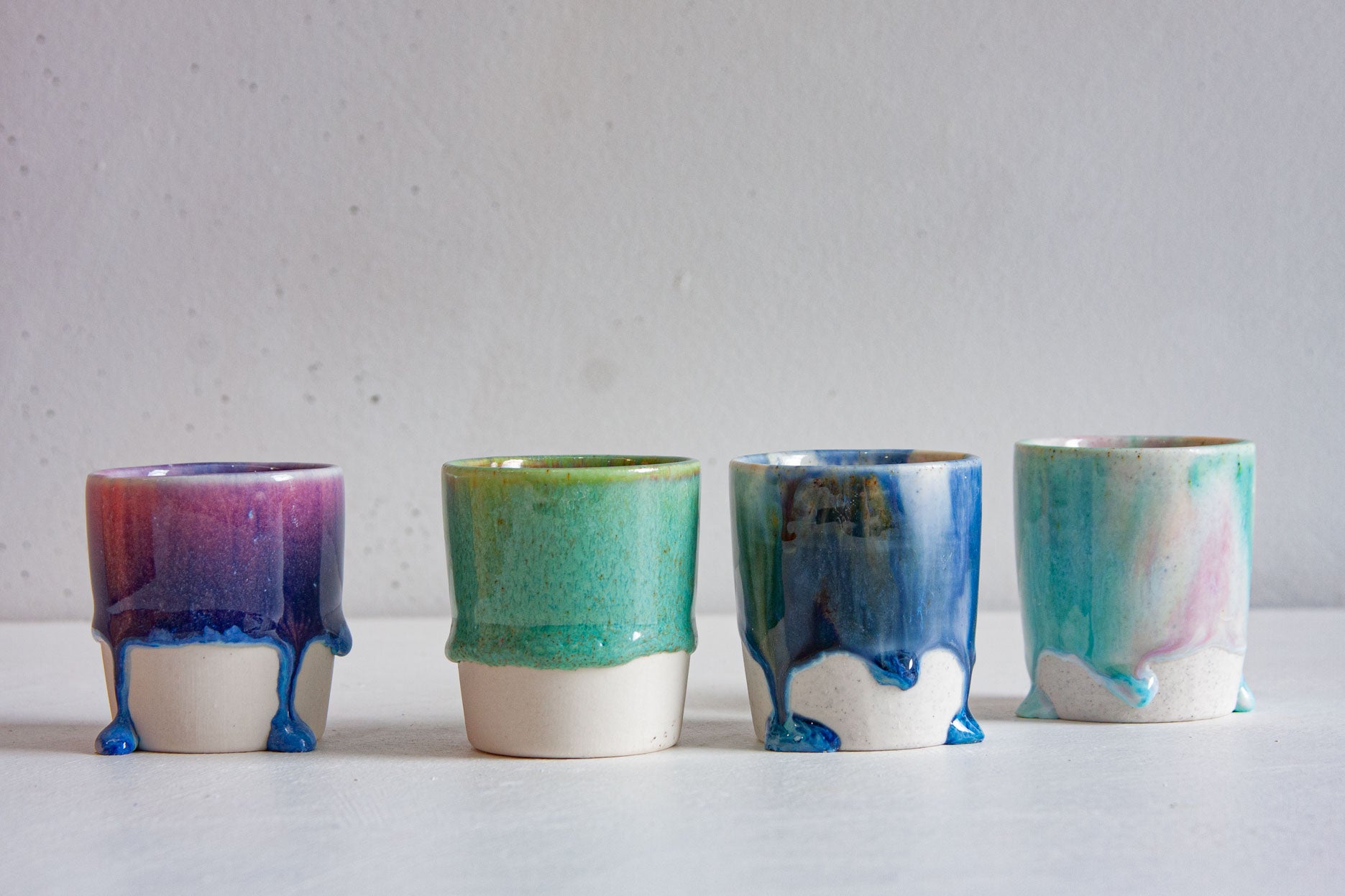 Four handmade ceramic cups in a row, each in a different glaze: purple fading into blue, jade green, deep blue with ochre, and soft mint with pink, all over bare clay bases.