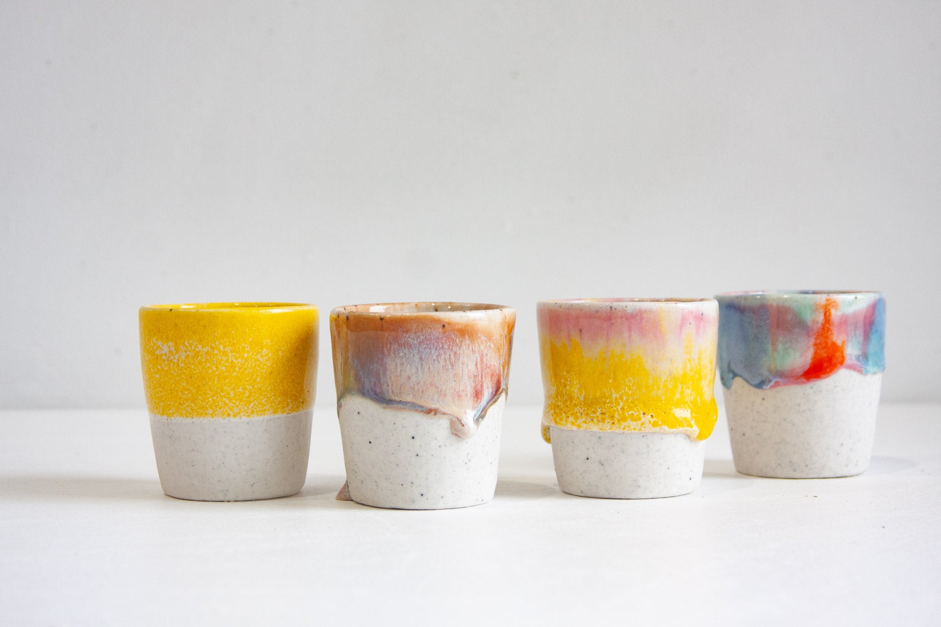 Four handmade ceramic espresso cups in a row, one solid yellow, one in peach and lilac, one yellow with pink at the rim and one in blue and teal with red, each on a speckled unglazed base.