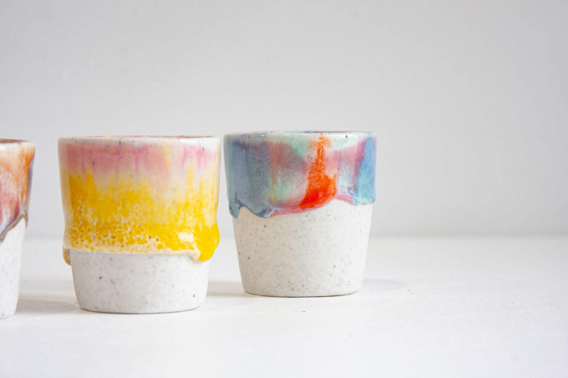 Two handmade ceramic espresso cups close up, one glazed yellow with pink above it, the other in blue and teal cut through with a run of red, both on heavily speckled unglazed bases.