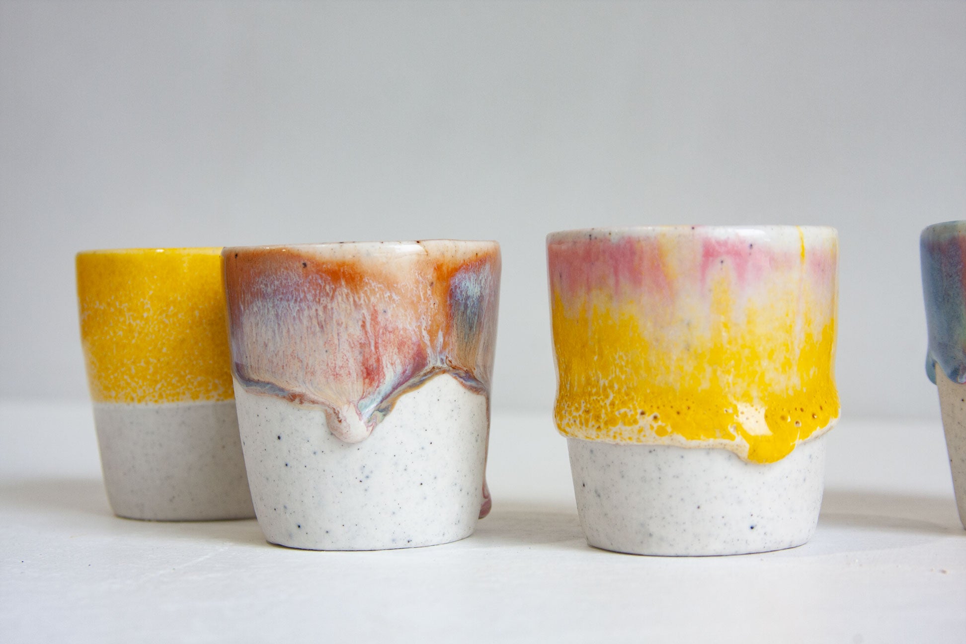 Three handmade ceramic espresso cups close up, in solid yellow, in peach and lilac fading to pink, and in yellow with pink at the rim, the glaze pooling thickly above the speckled bases.