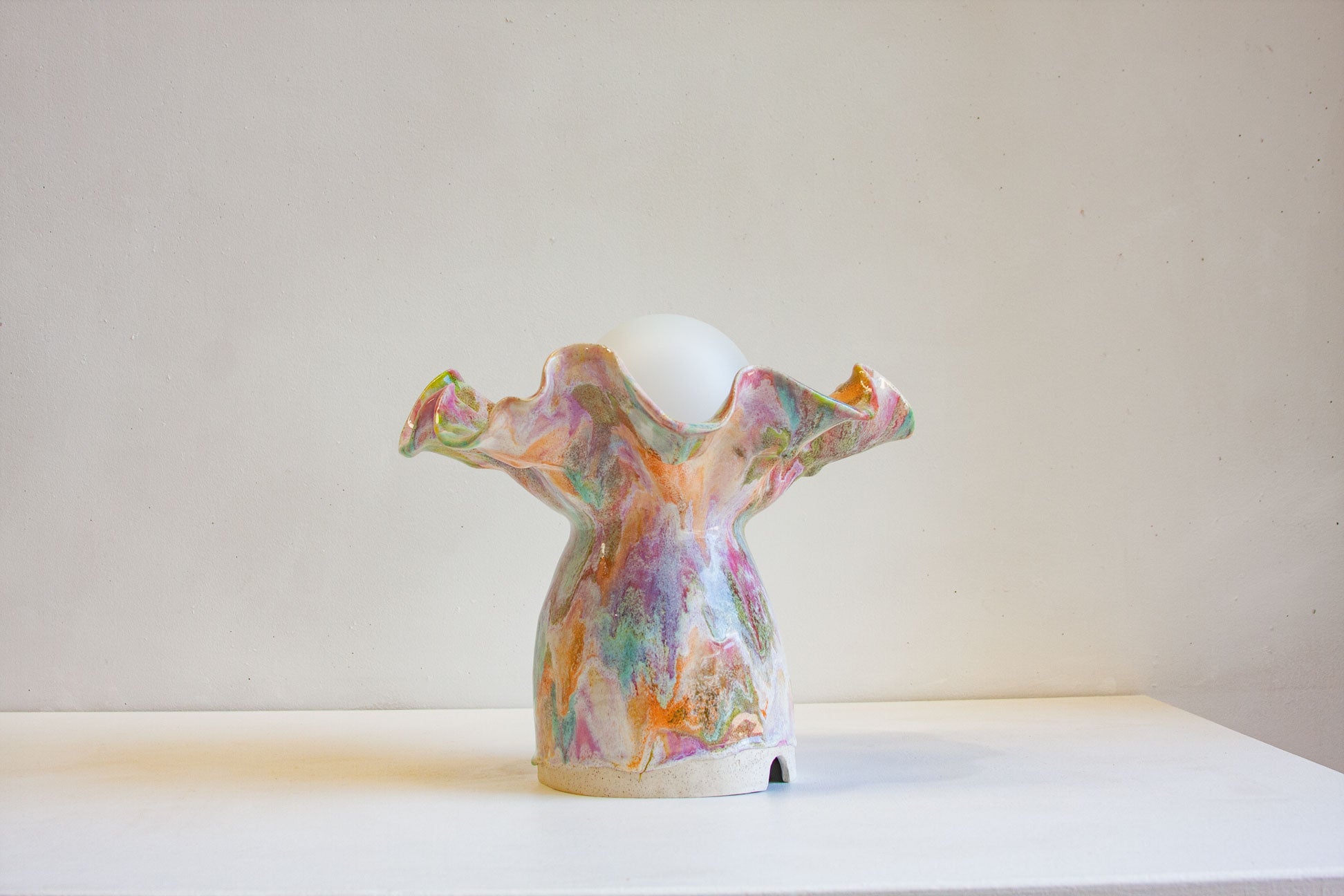 A sculptural ceramic table lamp with a ruffled, organic silhouette in pastel multicolor glaze, featuring a white spherical bulb at the top, displayed on a white surface against a neutral beige wall.