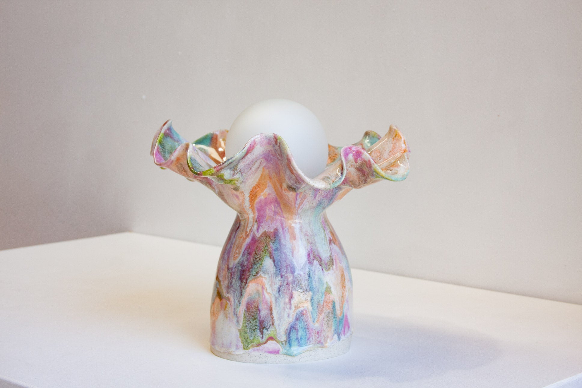 A handmade ceramic vase with a colorful marbled pattern in pink, purple, green, and orange, holding a white egg-shaped object on a white surface against a neutral gray background.